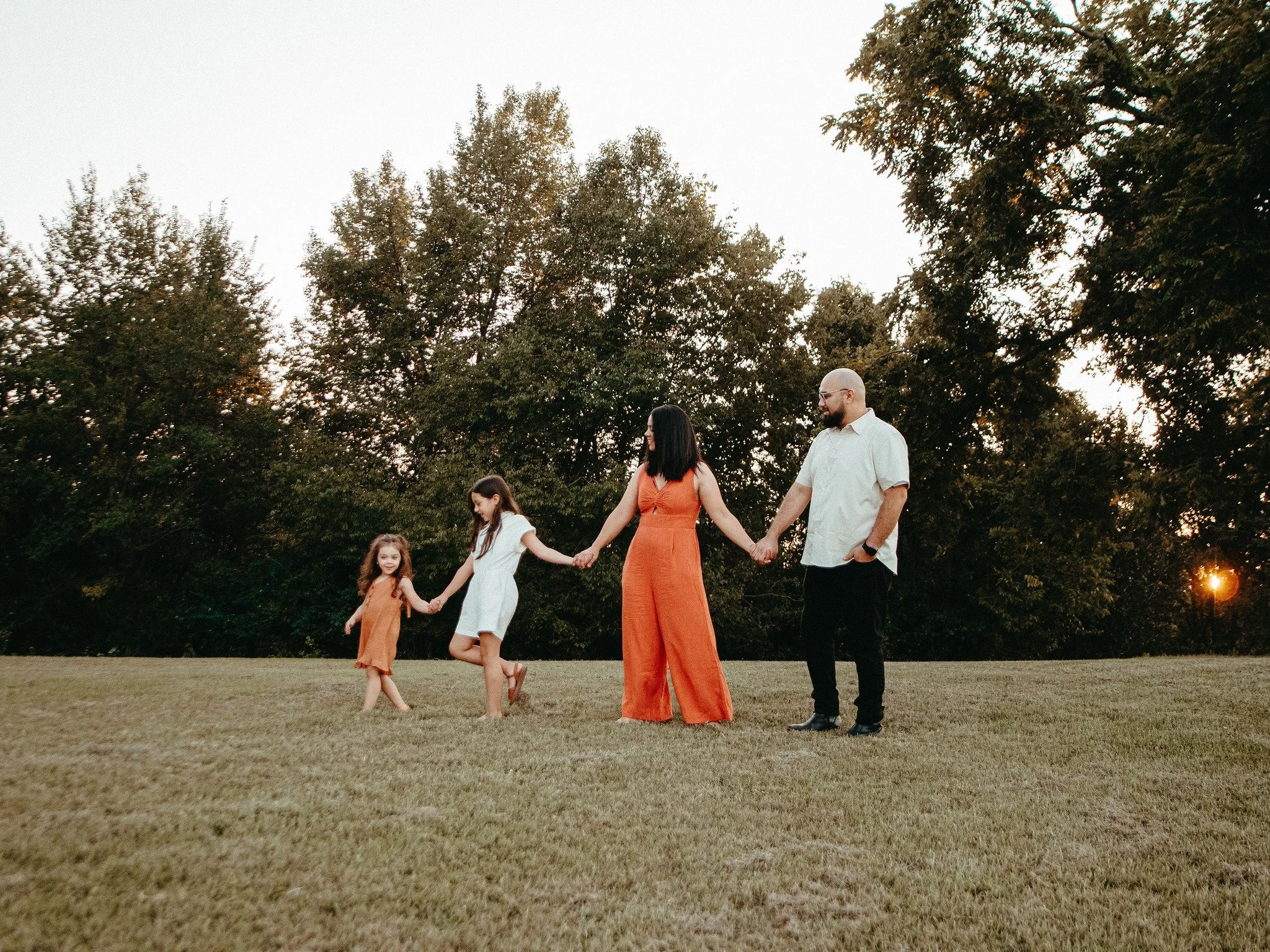 Family of four holding hands and walking across a grassy field during sunset, with trees in the background.