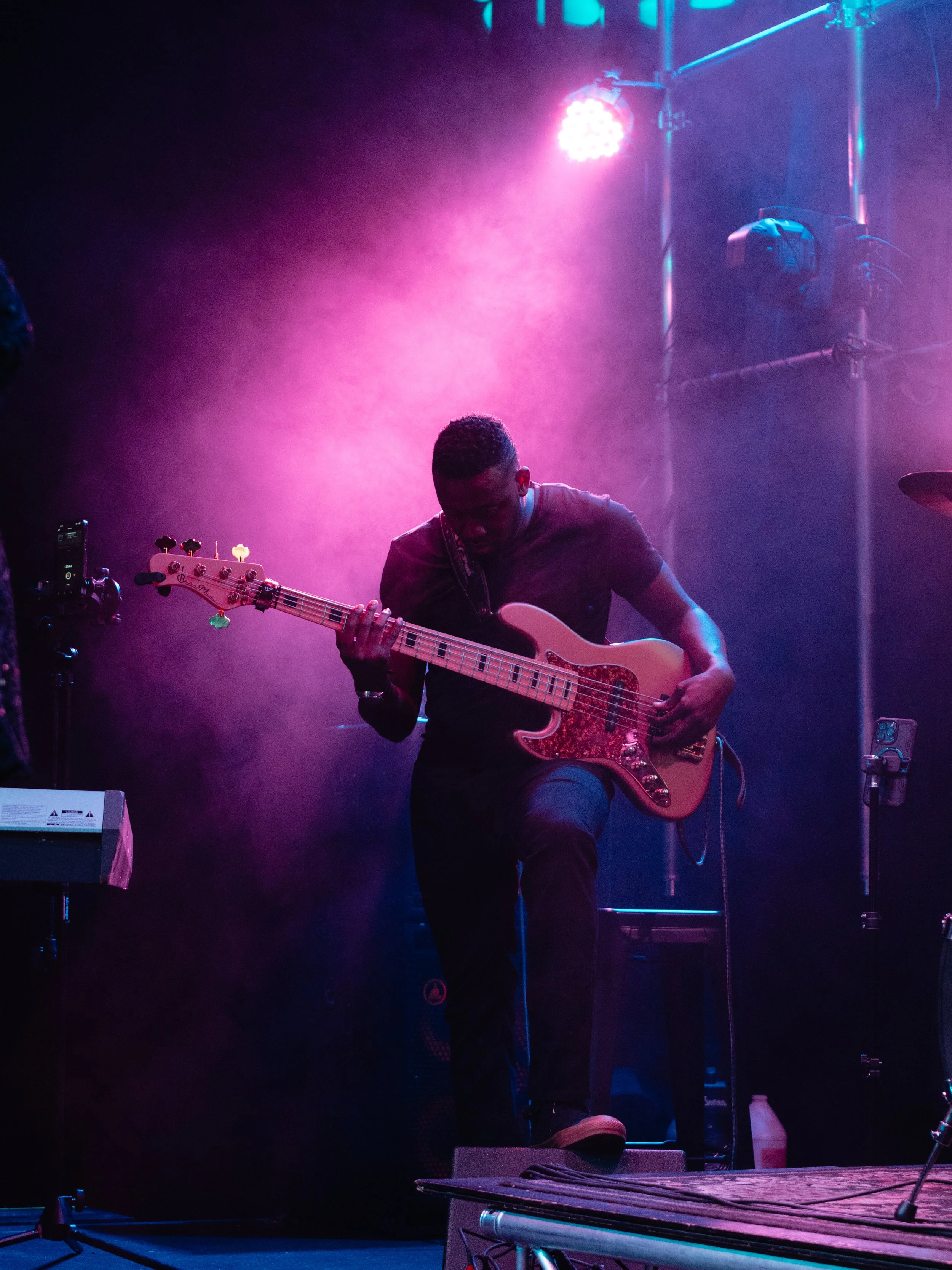 A musician playing an electric bass guitar on stage under colorful pink and blue lighting with fog effects.