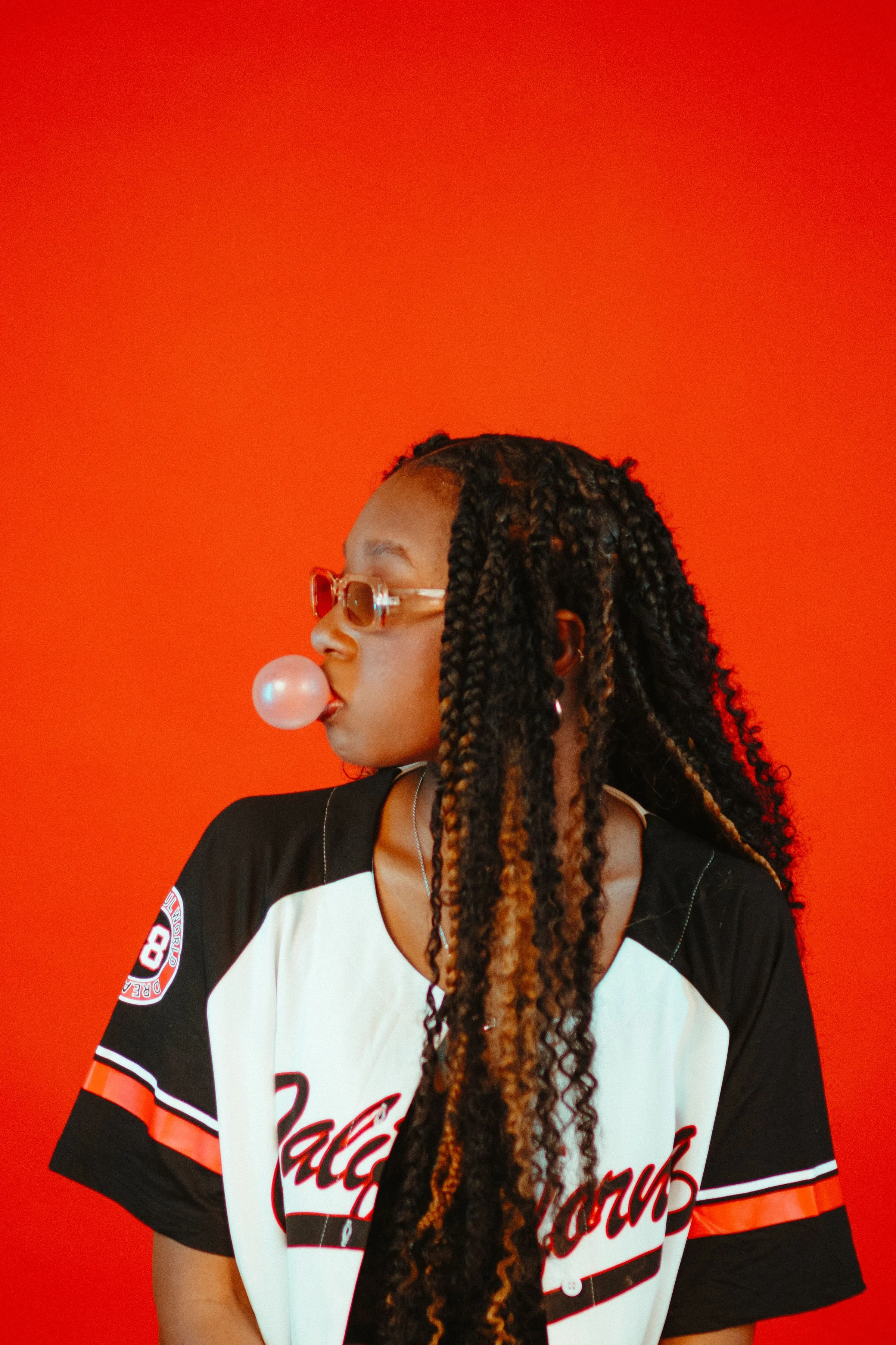 Woman with long braided hair wearing sunglasses blowing pink bubble gum against an orange background.