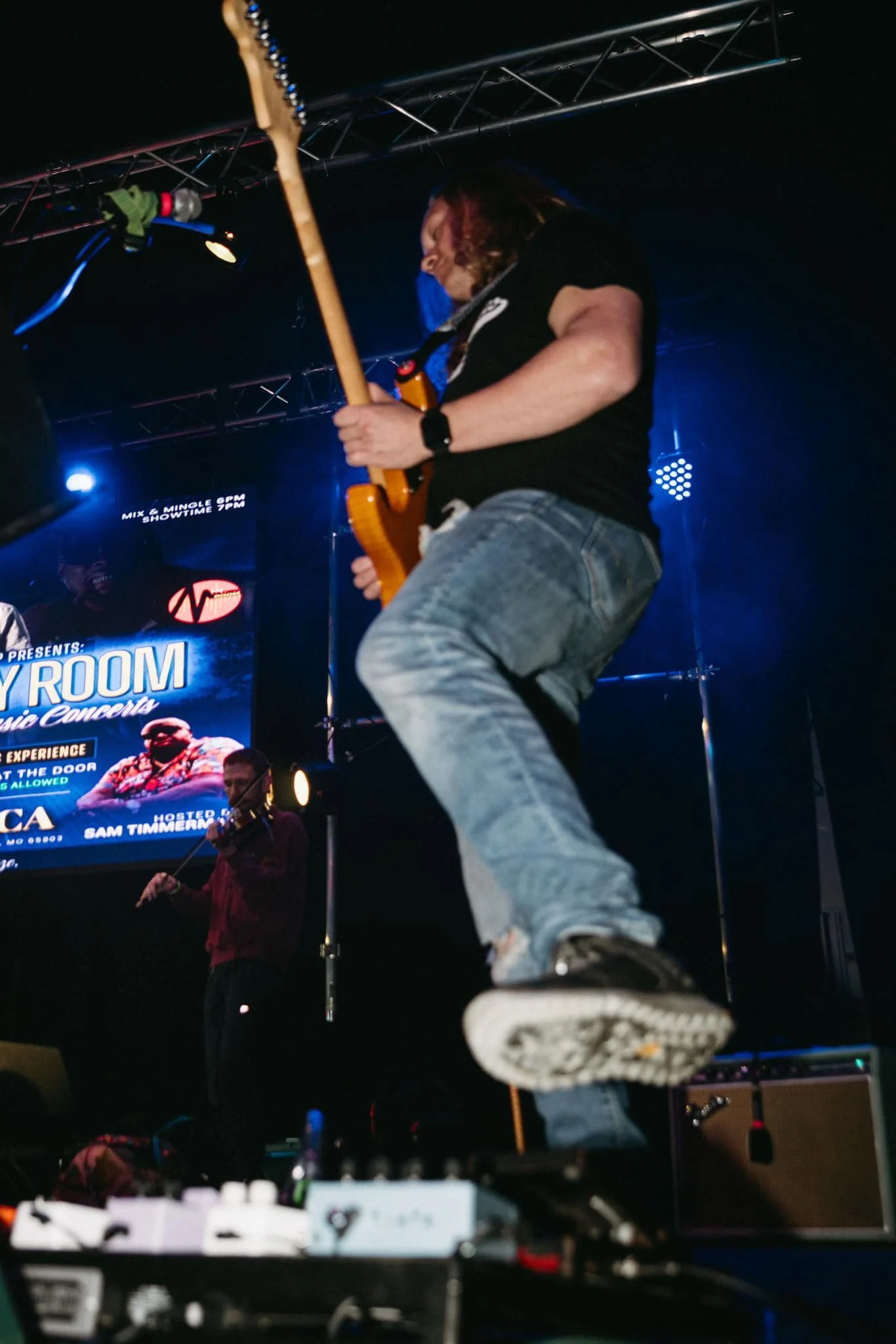Guitarist jumping on stage during a concert with a screen in the background displaying event details.
