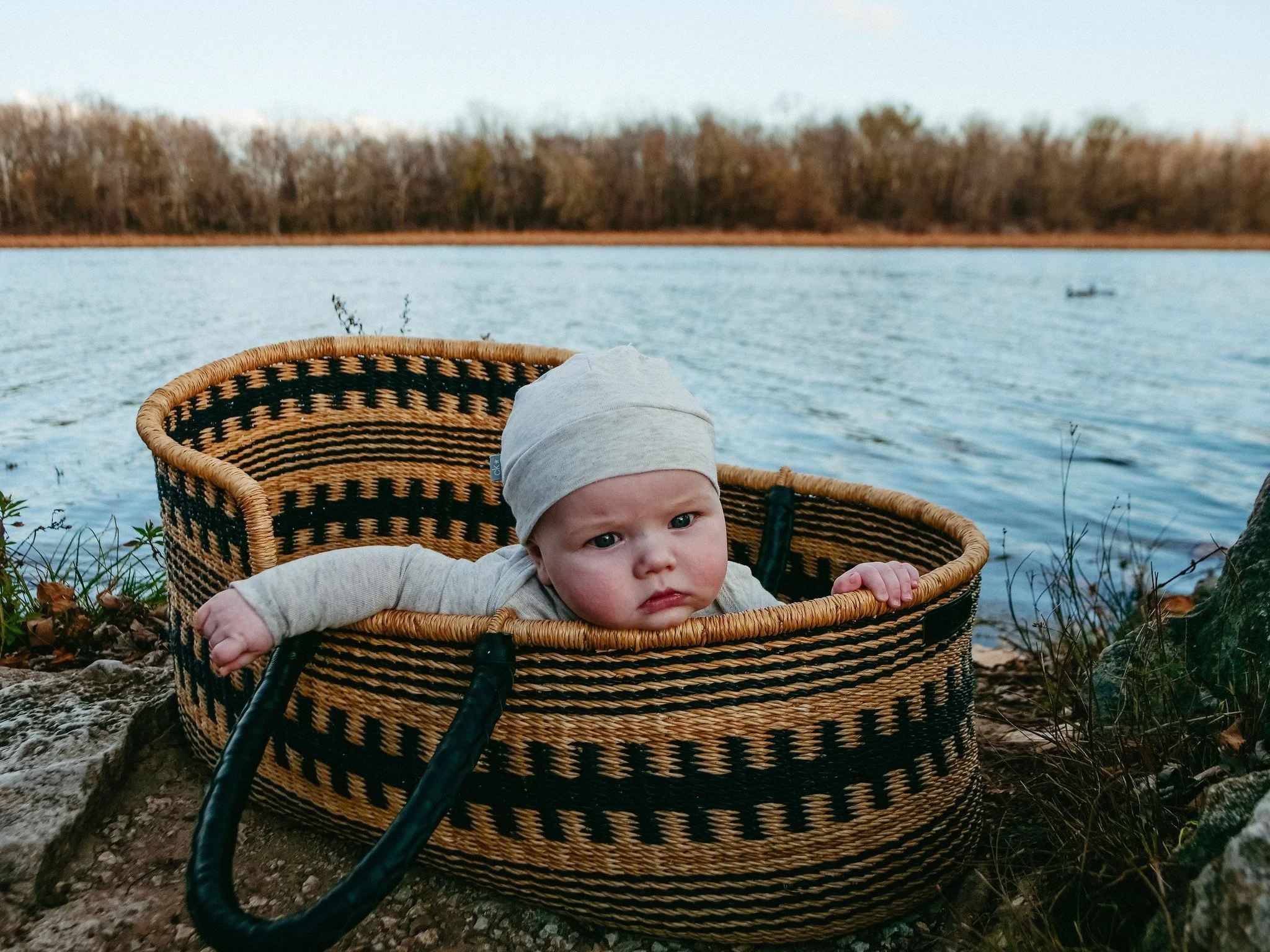 A baby with a gray hat and beige clothing resting in a patterned woven basket by a lake, with trees in the background and a small boat on the water.