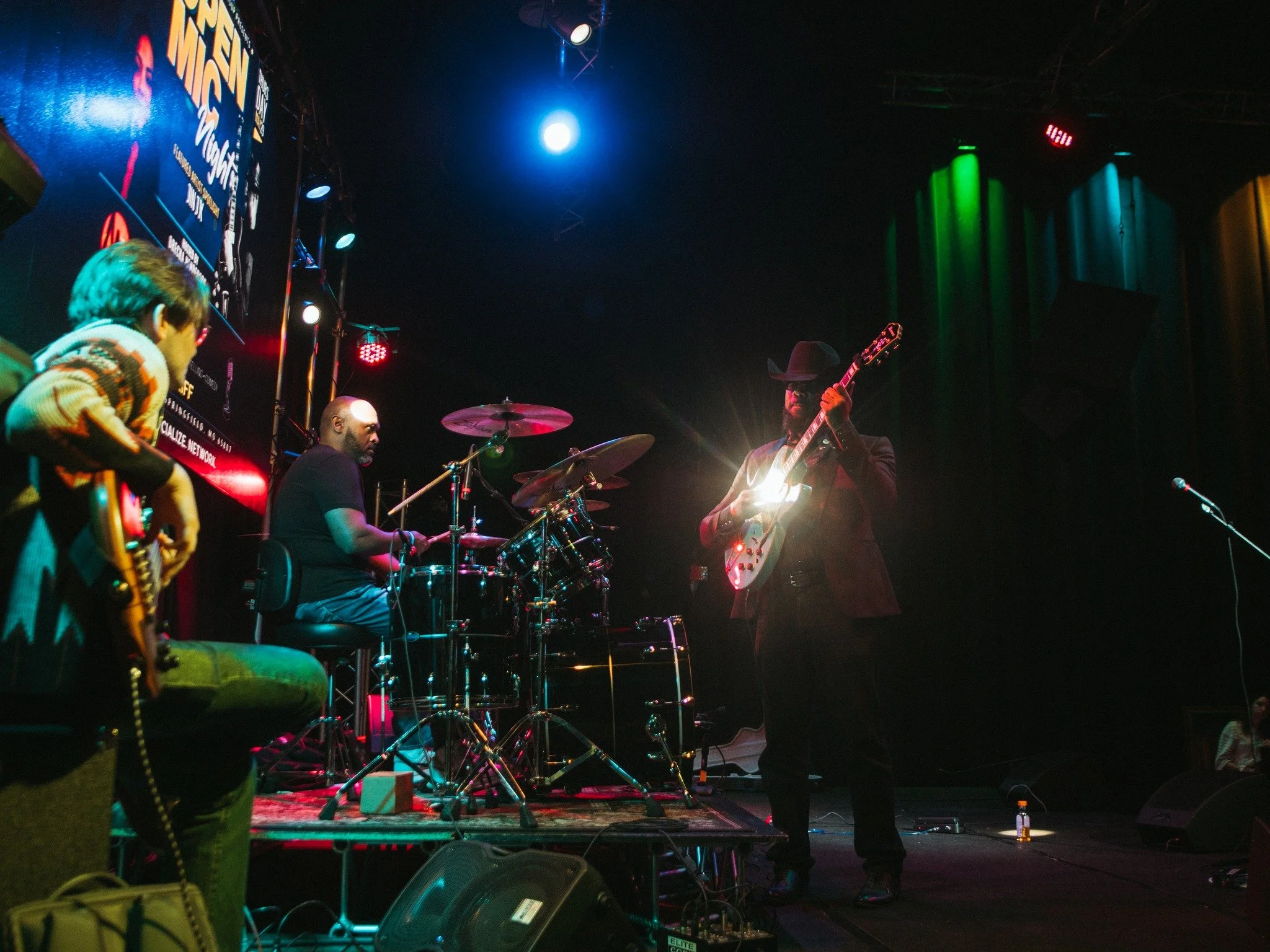 Musicians performing on a stage with colorful lighting, including a guitarist in a cowboy hat and a drummer, during a concert.