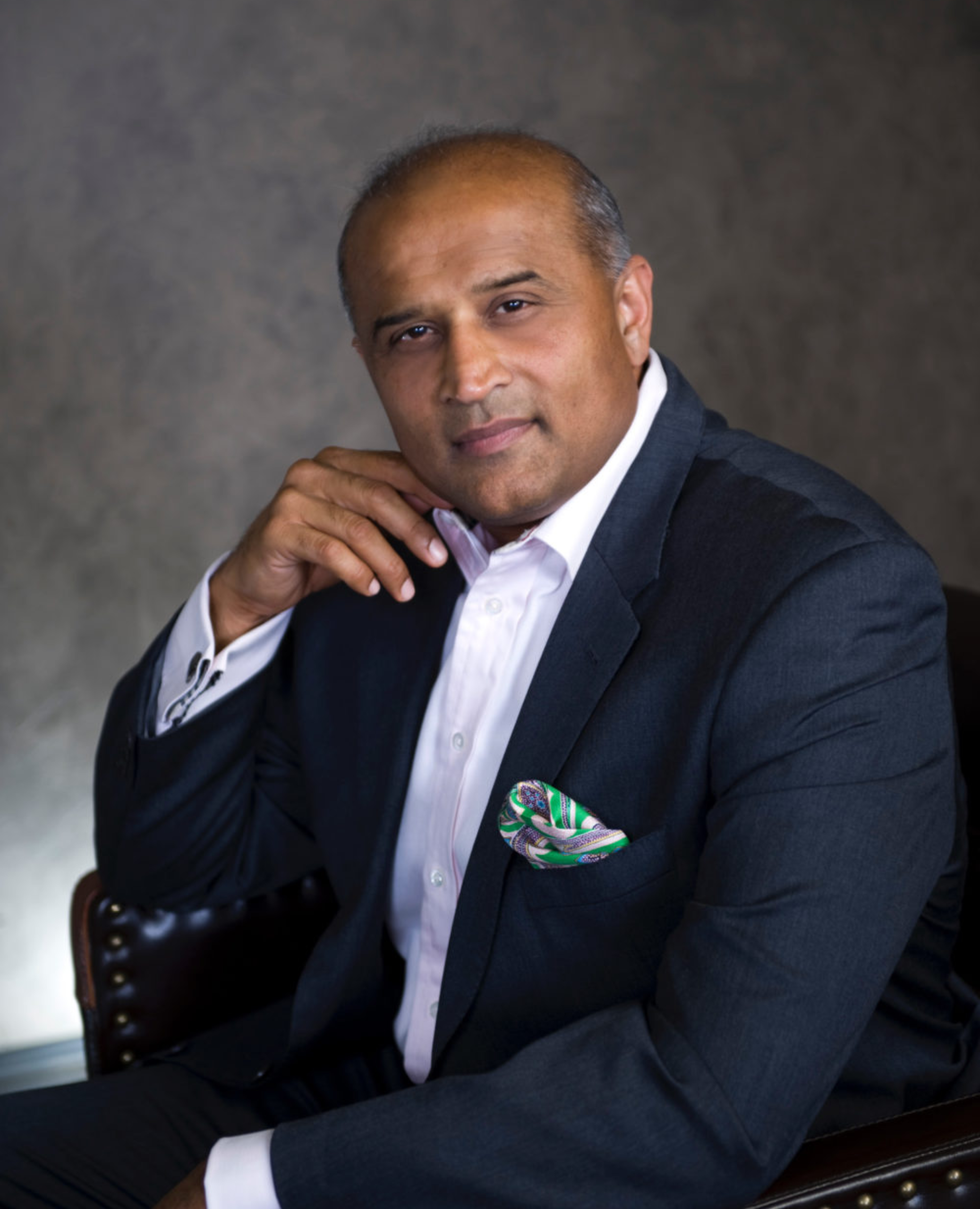 A man in a dark suit, white shirt, with a colorful pocket square, sitting on a chair, with his hand resting near his face, looking at the camera.