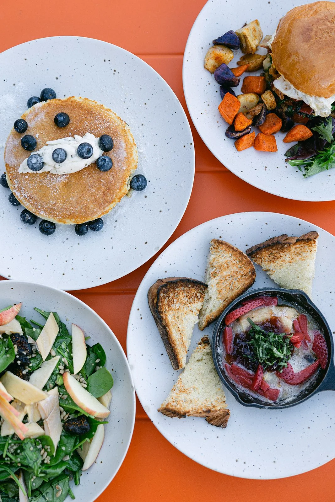 Assorted dishes including blueberry pancakes, veggie burger with sweet potato fries, salad with apples, and bread with dip on orange table.