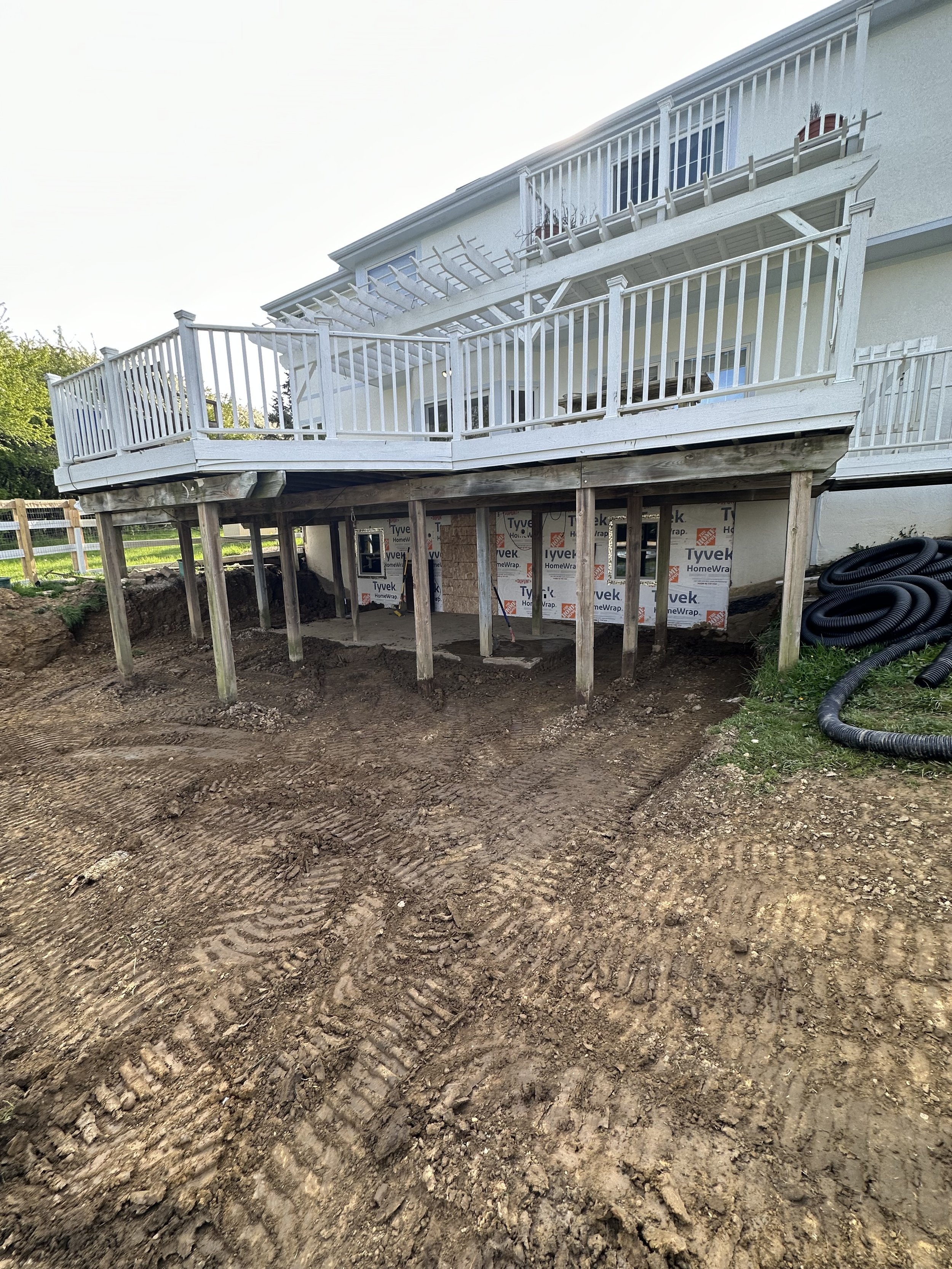 Construction site showing a house with a balcony, the area underneath being prepared with posts and dirt for further work.