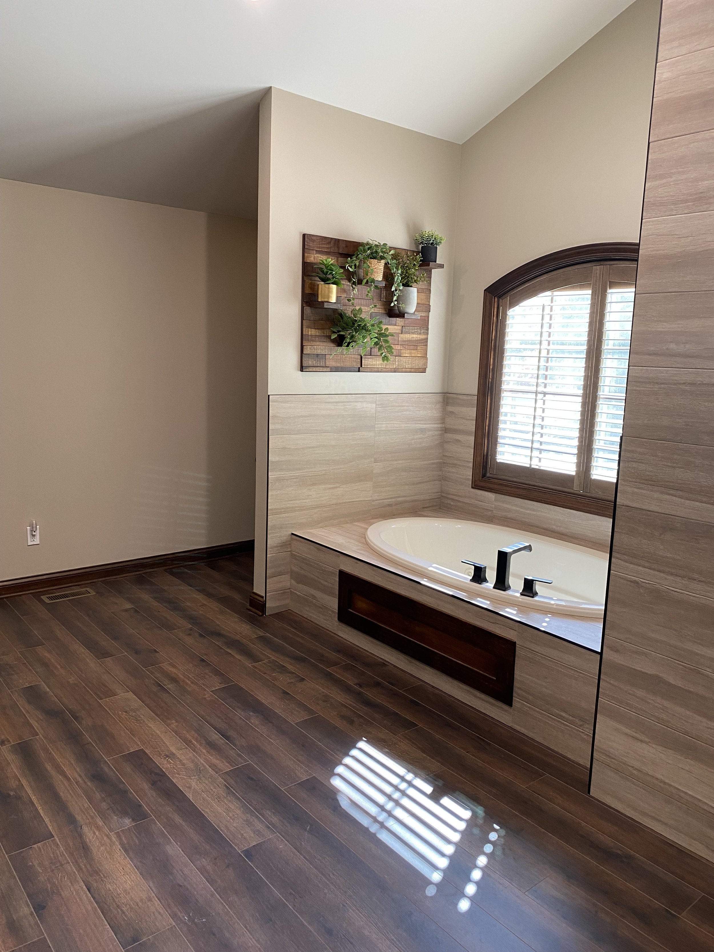 Bathroom with a bathtub beneath a window with wooden blinds, a wall-mounted wooden planter with green plants, and a wooden frame mirror reflecting the room.