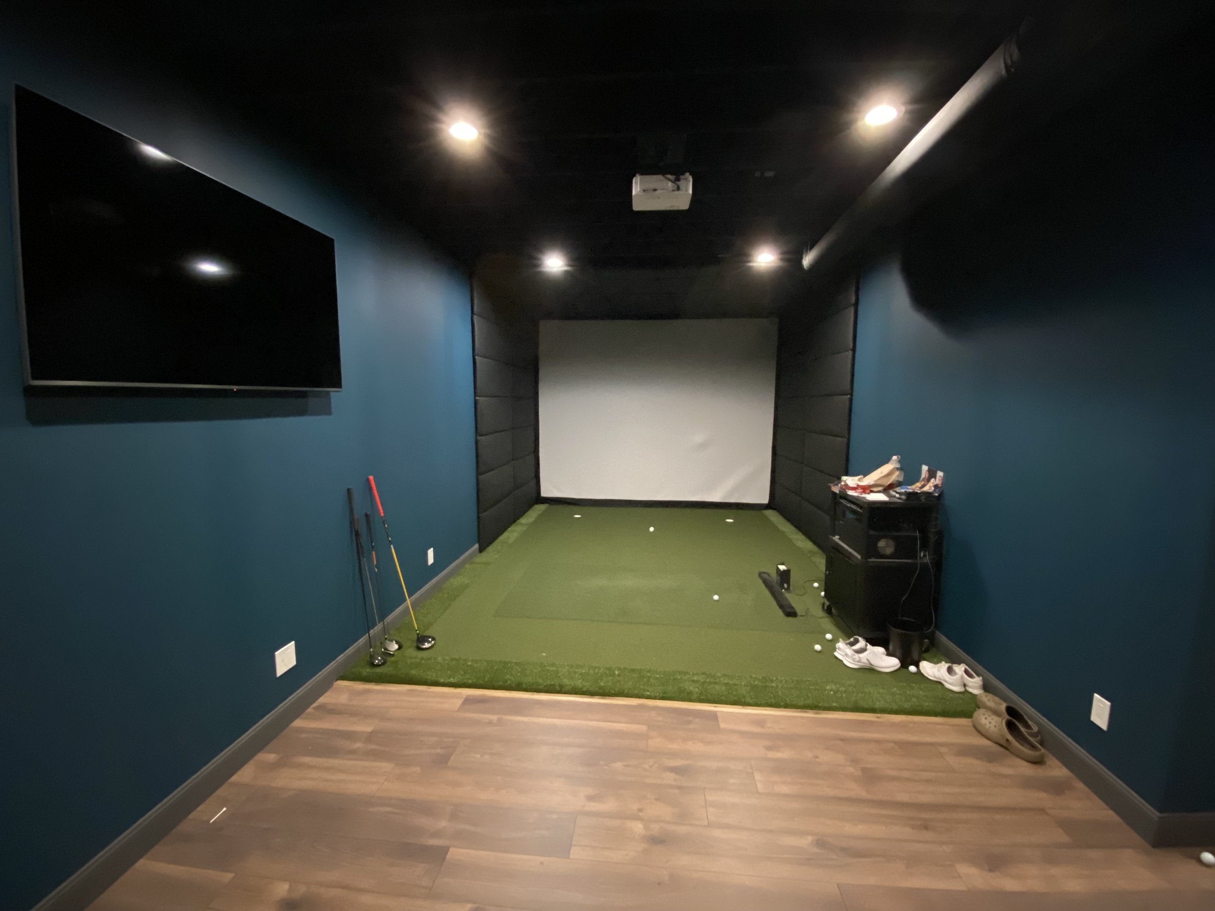 Indoor golf simulation room with a grass mat, golf clubs, golf balls, and a large screen for virtual golf games.