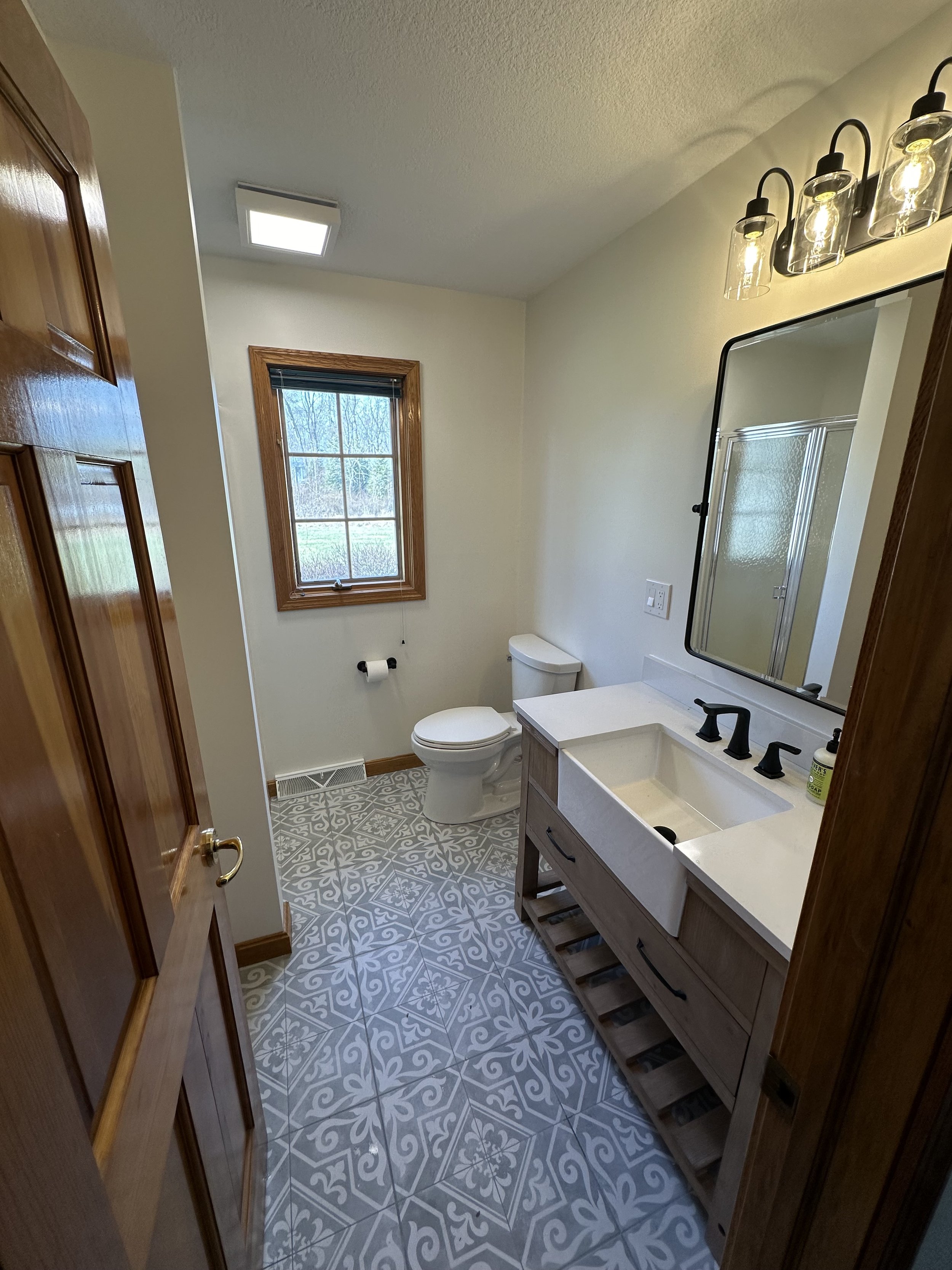 Bathroom with a white toilet, wooden-framed window, vanity with a farmhouse sink, mirror, black fixtures, and patterned tile flooring.