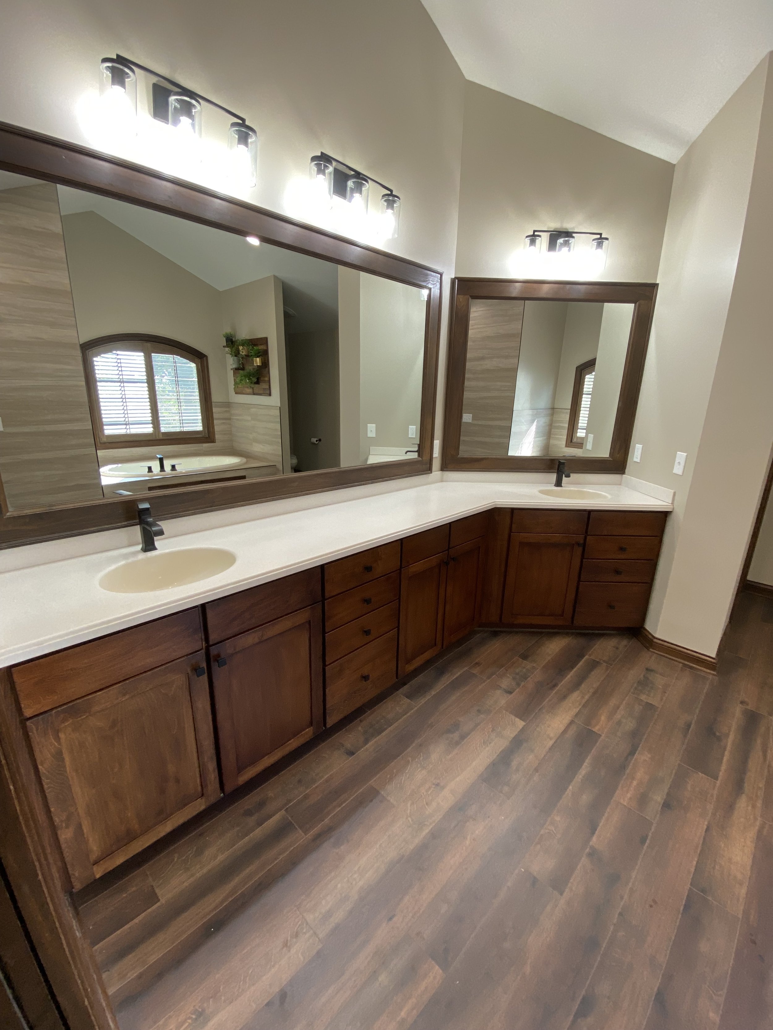 A bathroom with a double wooden vanity, large mirrors, and wood flooring. There are two sinks with black faucets, and above each mirror are two light fixtures with multiple bulbs. The bathroom has beige walls, and a window can be seen in the reflecti