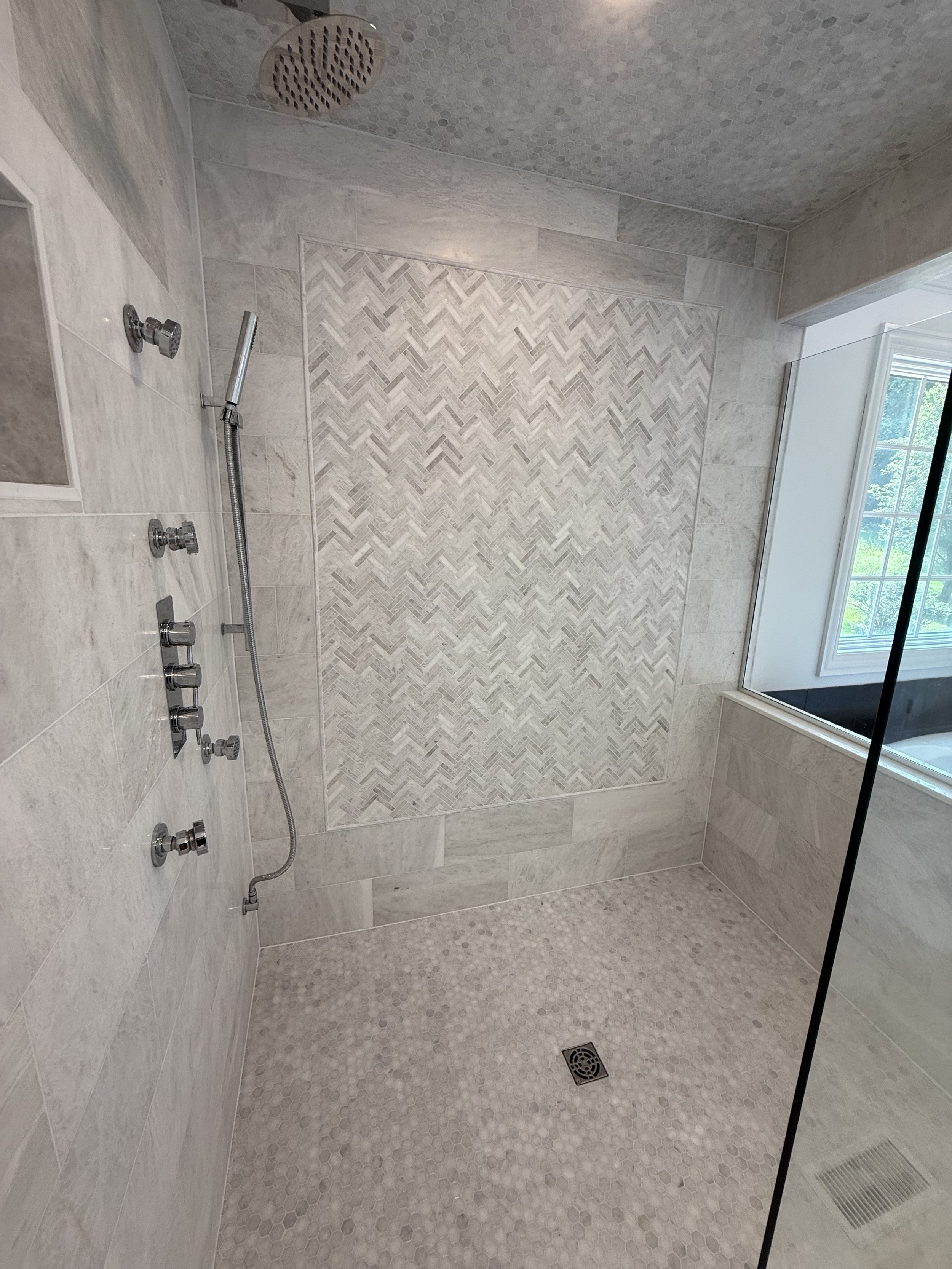 A modern walk-in shower with light-colored tile walls, a textured mosaic tile accent wall, and a matching mosaic tile floor. There is a window to the right and a ceiling-mounted showerhead.