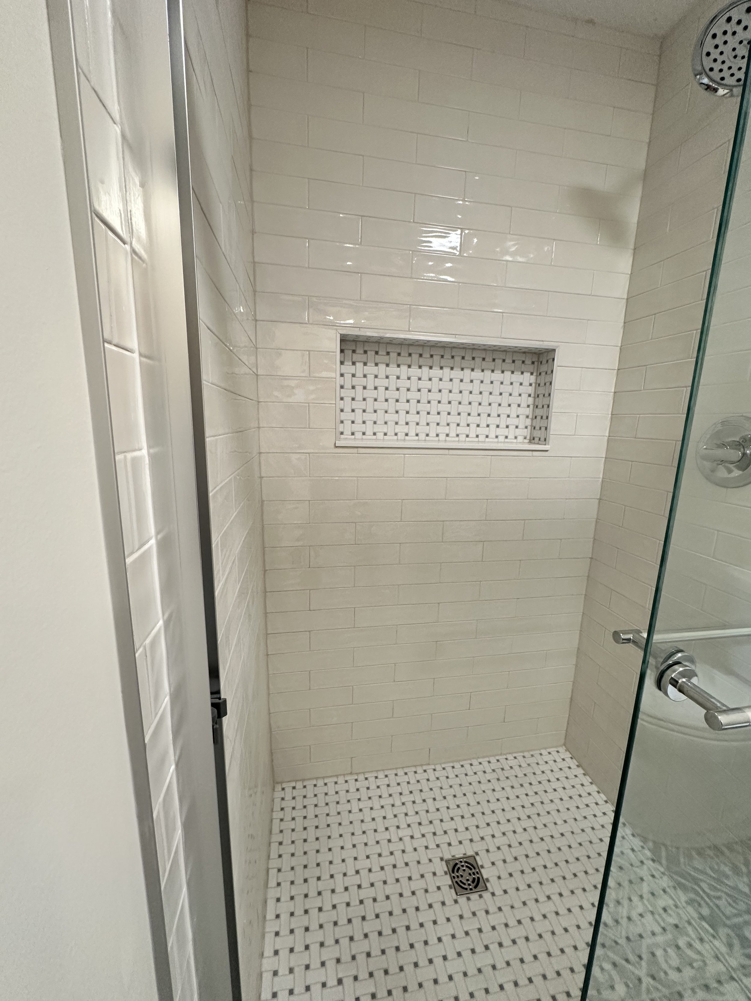 Inside a modern shower with cream-colored subway tiles, a built-in basket niche, and a white herringbone patterned tile floor with a drain in the center.