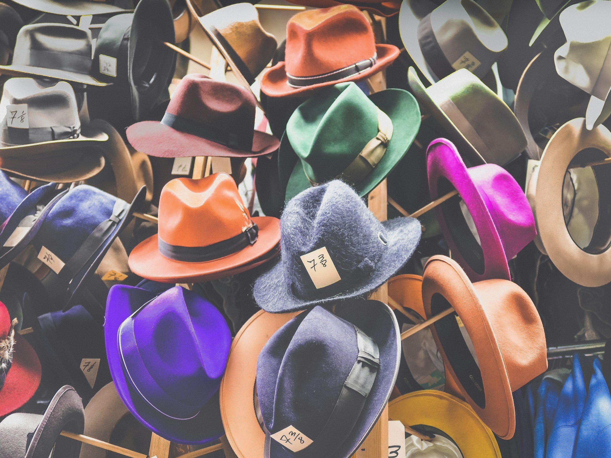 A variety of colorful hats including fedoras and wide-brim hats on display with price tags, arranged on a multi-tiered stand.