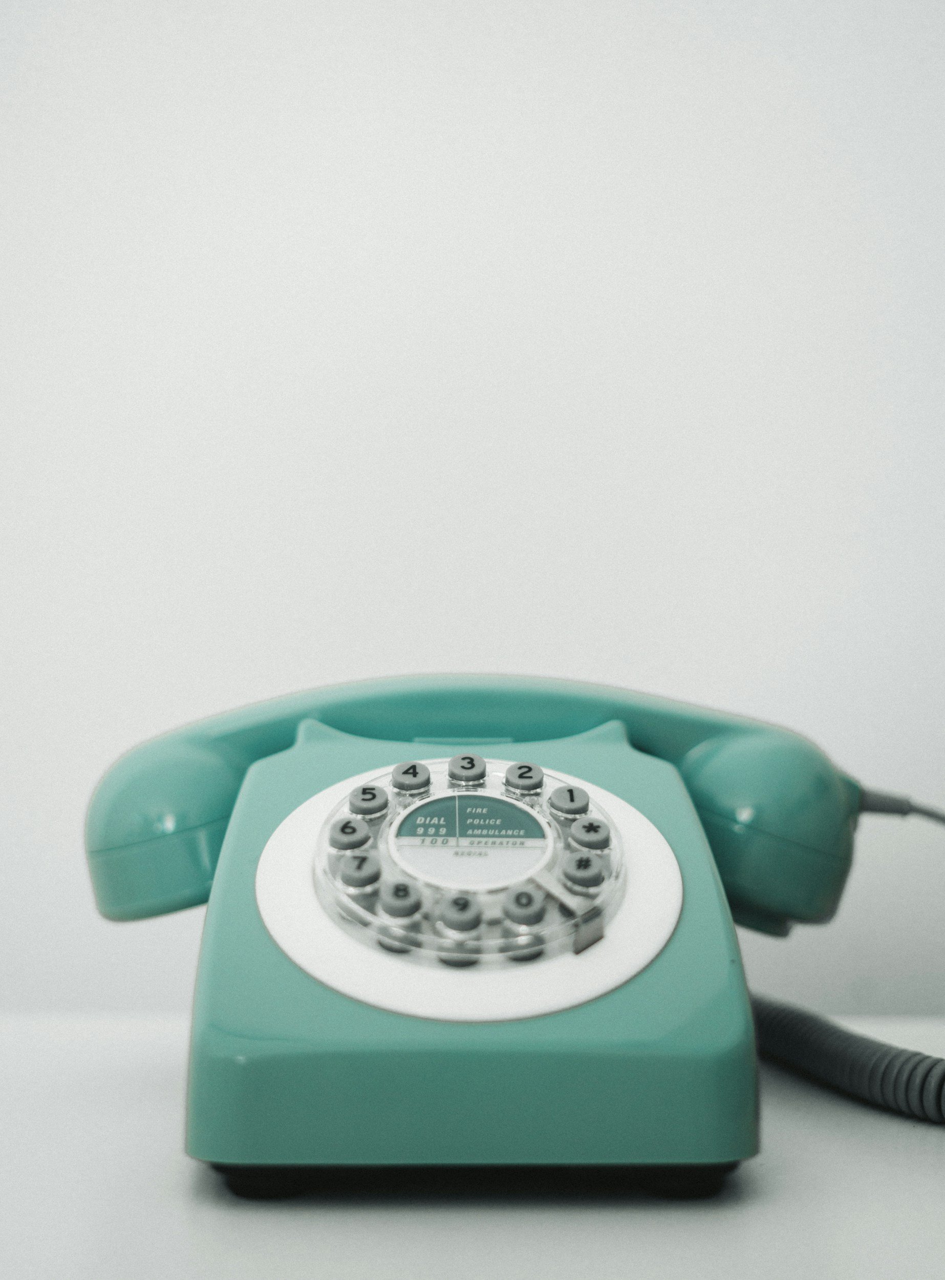 A retro teal rotary telephone with a coiled cord, sitting on a plain white surface.