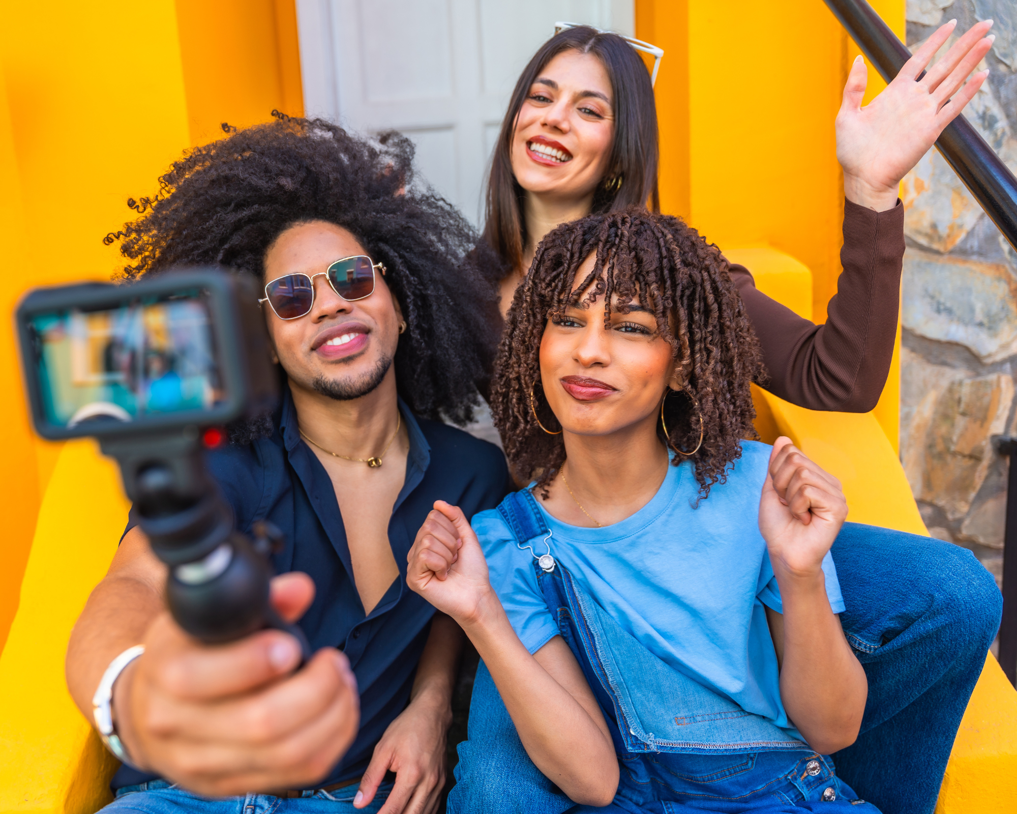 Group of four young adults taking a selfie outdoors with a smartphone, smiling and making gestures, against a vibrant yellow wall.