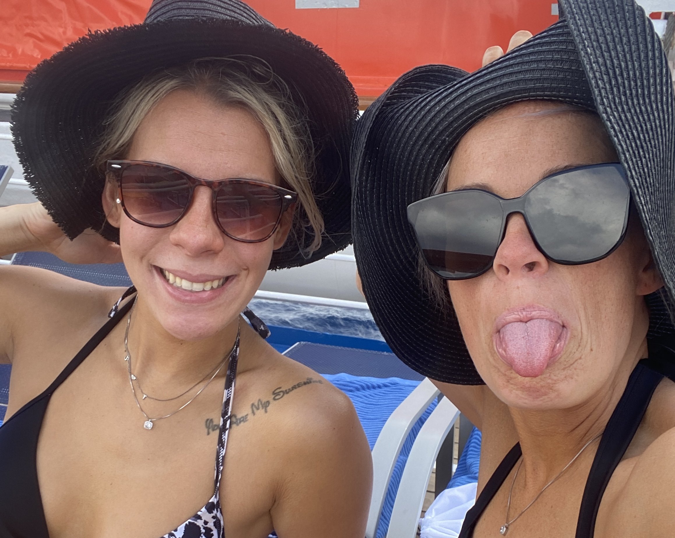 Two women wearing large black sun hats and sunglasses, smiling and sticking out their tongues while relaxing outdoors on a boat or poolside.