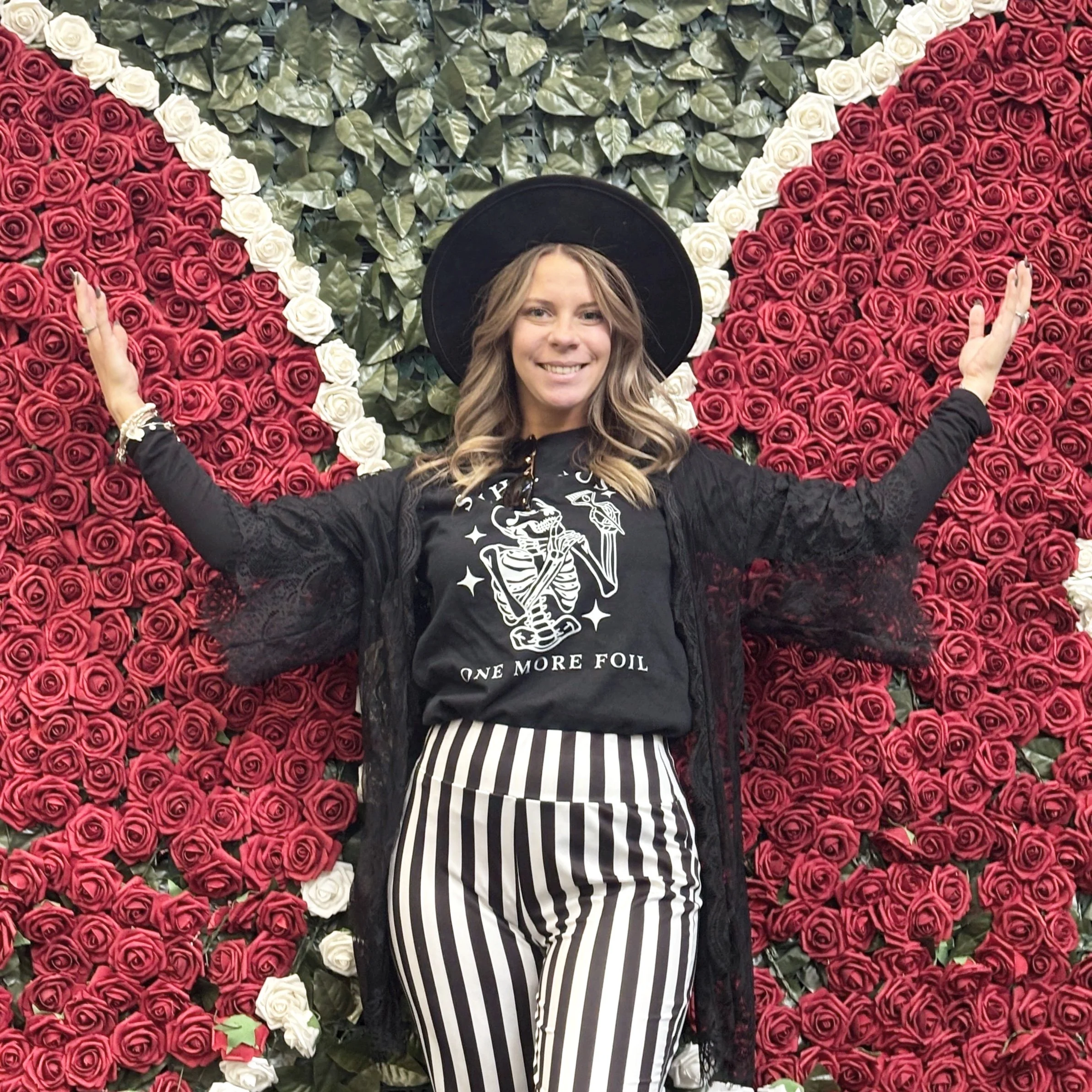 Woman wearing a black wide-brimmed hat, black lace cover-up, striped black and white pants, and a black T-shirt with a skeleton and the phrase 'One More Foil,' standing in front of a large wall of red and white roses shaped like a heart.