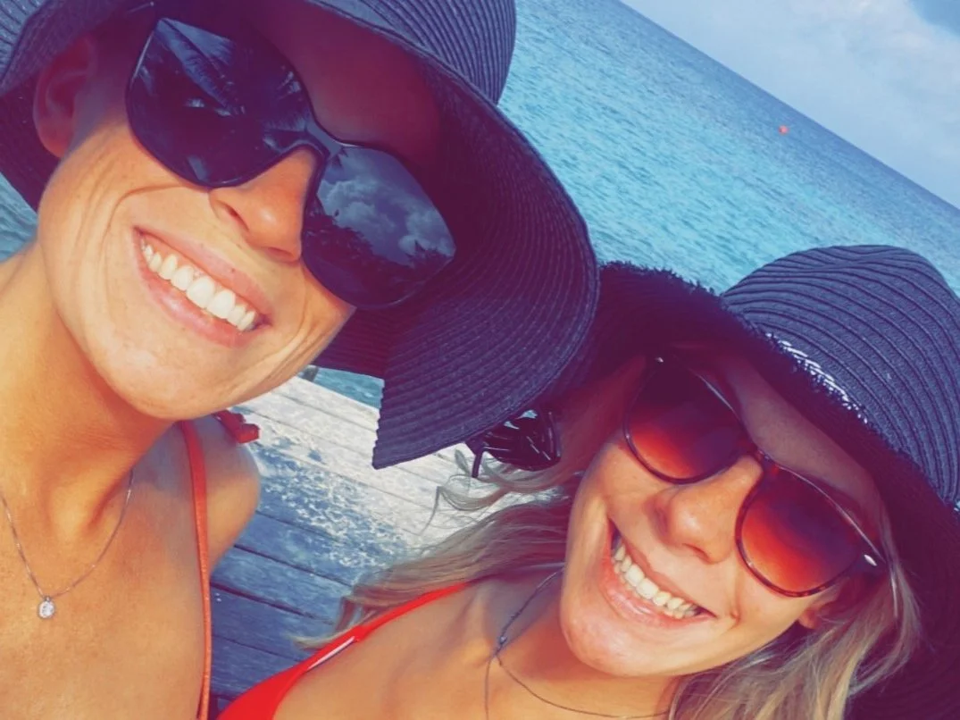 Two smiling women wearing large sun hats and sunglasses taking a selfie on a dock by the ocean.