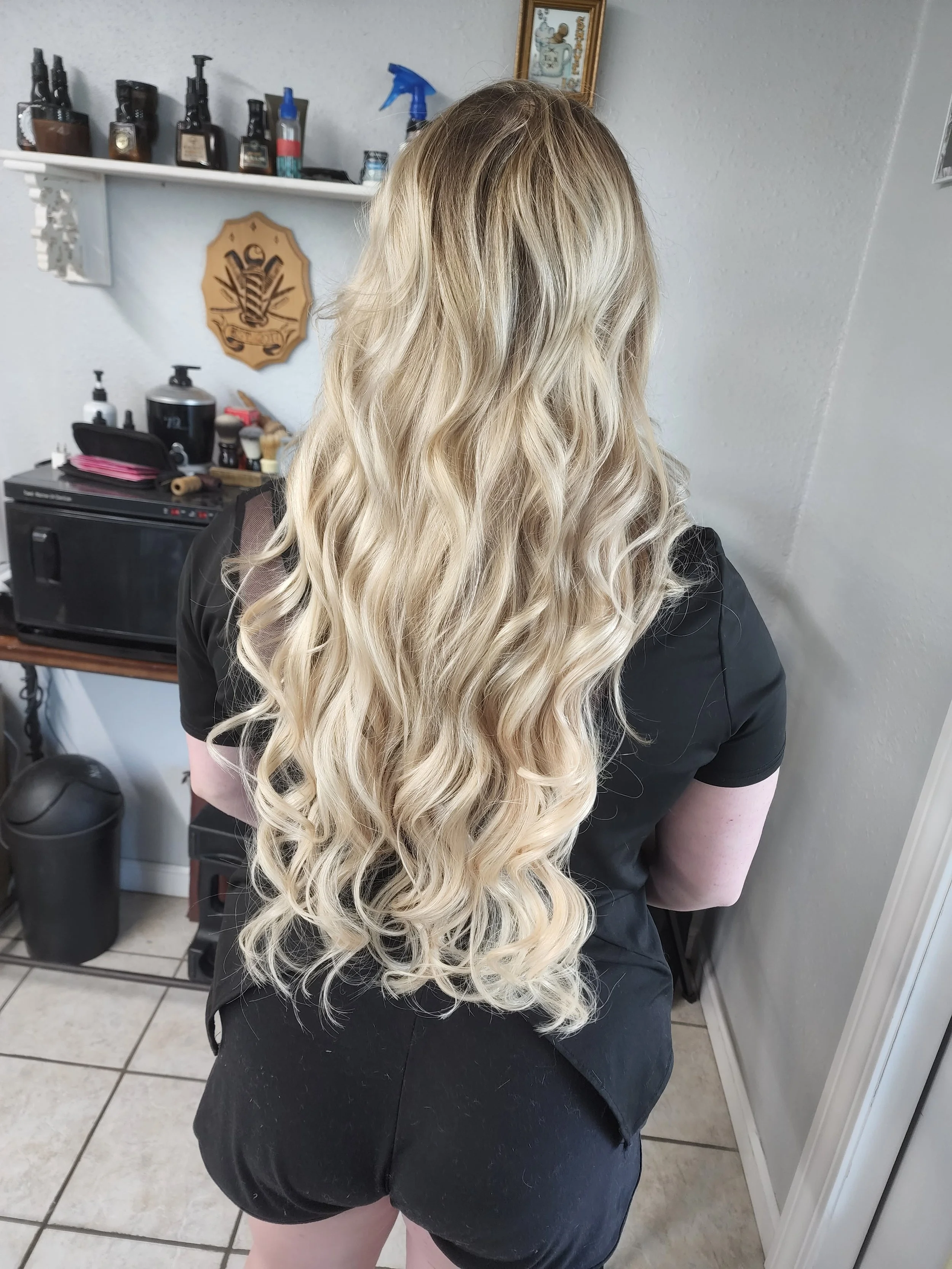 Back view of a woman with long, wavy blonde hair in a salon or home setting.