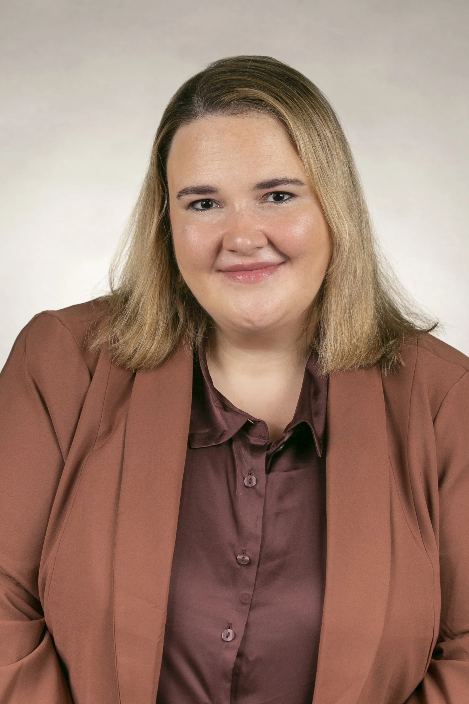 A woman with shoulder-length blonde hair, smiling, wearing a brown blazer and a maroon blouse, against a plain light-colored background.