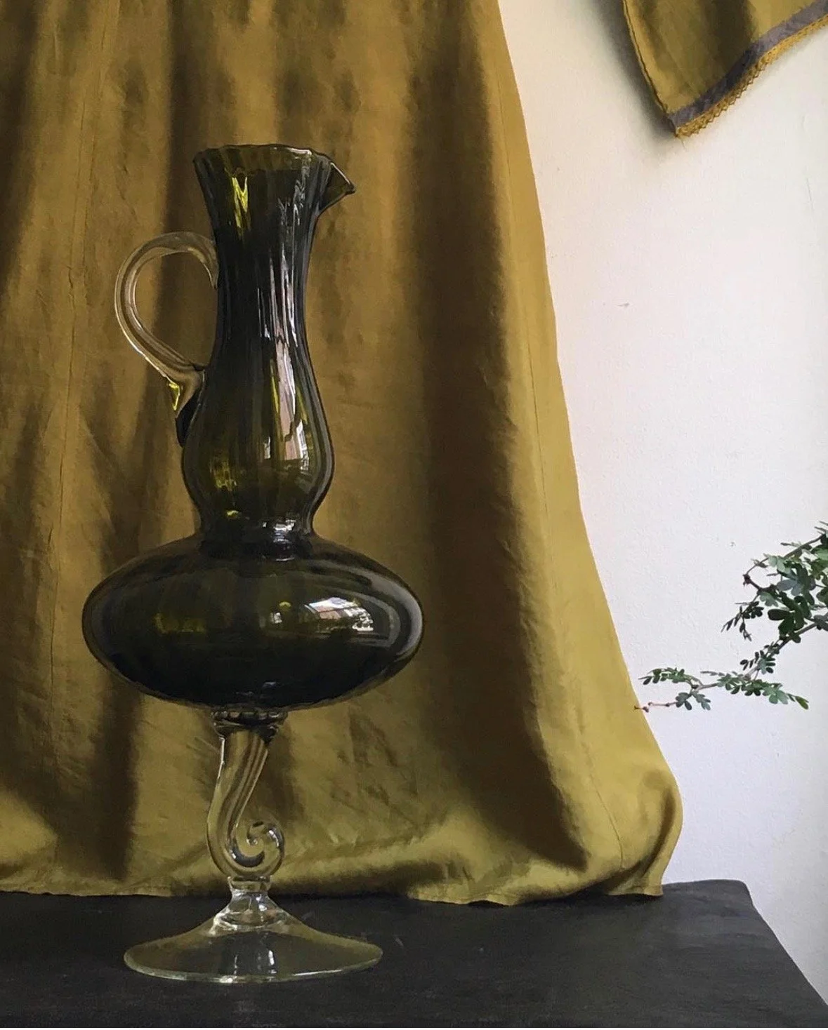 A glass vase with a black hue, featuring a narrow neck, a wide rounded base, and a spiral design on the stem, sitting on a black surface against a yellow curtain background with a small plant partially visible on the right.