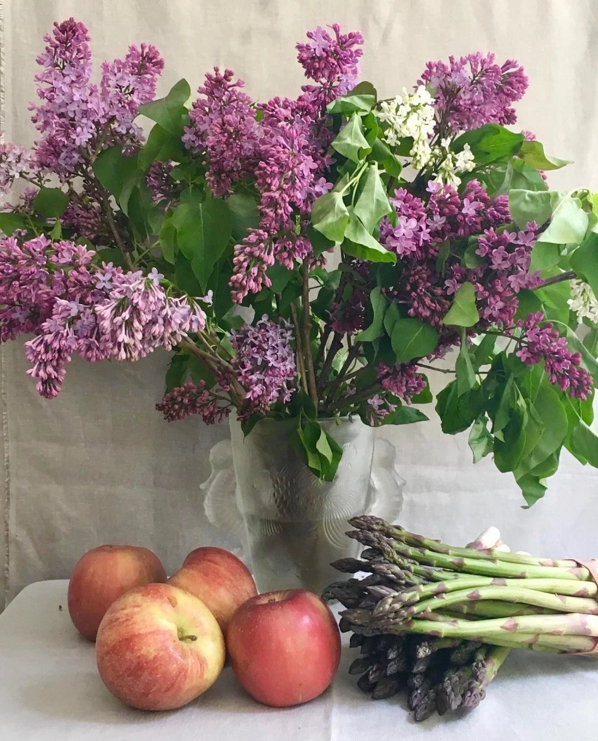 A vase with lilac flowers and green leaves, with five apples and a bunch of fresh asparagus on a table.