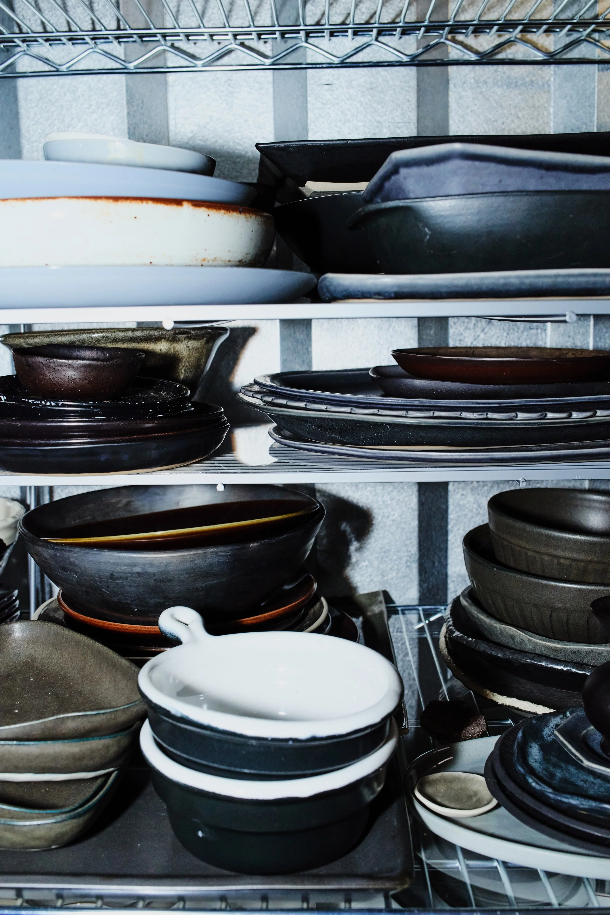 Stacked assorted ceramic and glass dinner plates and bowls on metal shelves in a kitchen storage area.