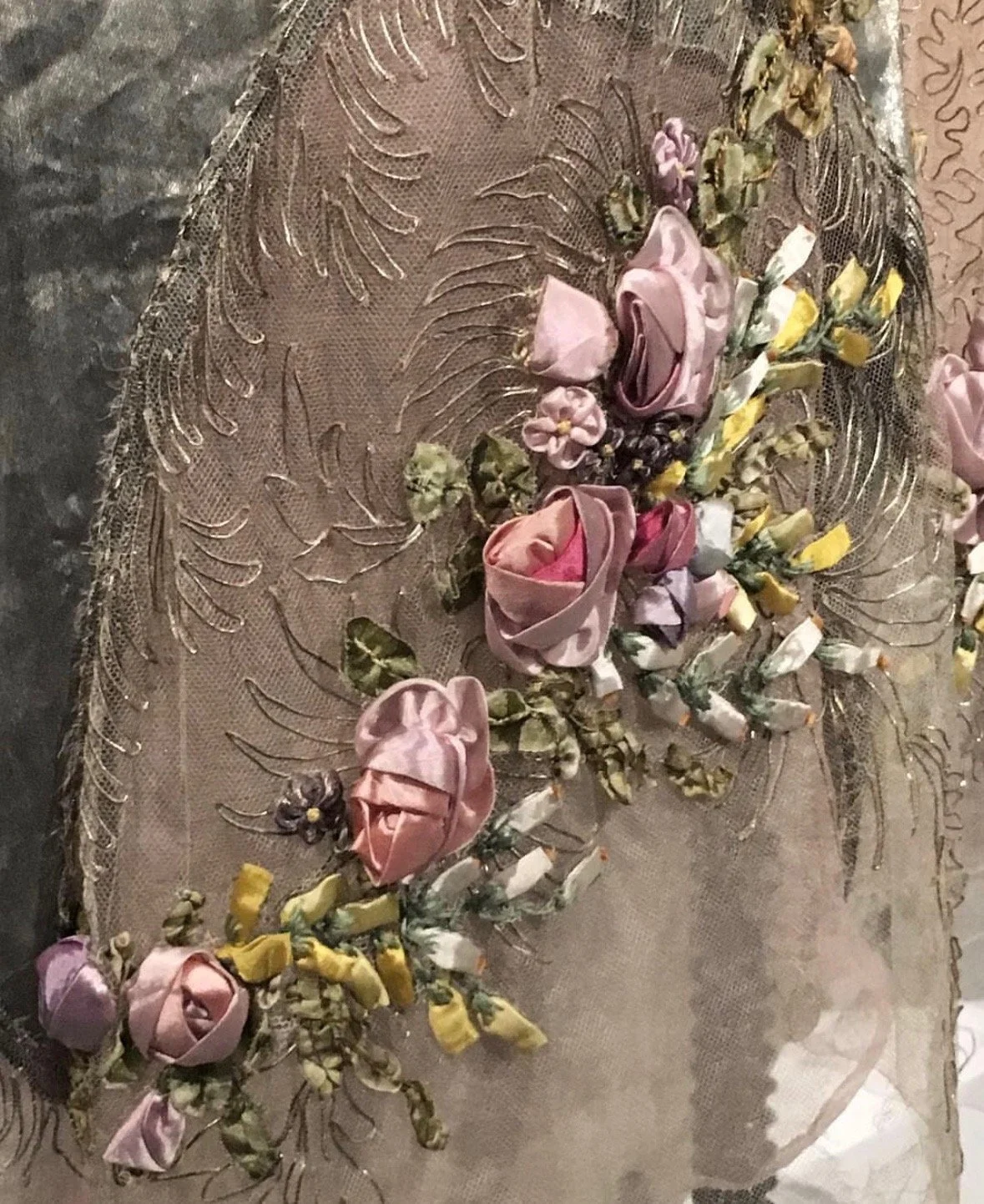 Close-up of a decorative fabric with embroidered floral details and silk flowers sewn onto it, featuring pink roses and yellow accents.