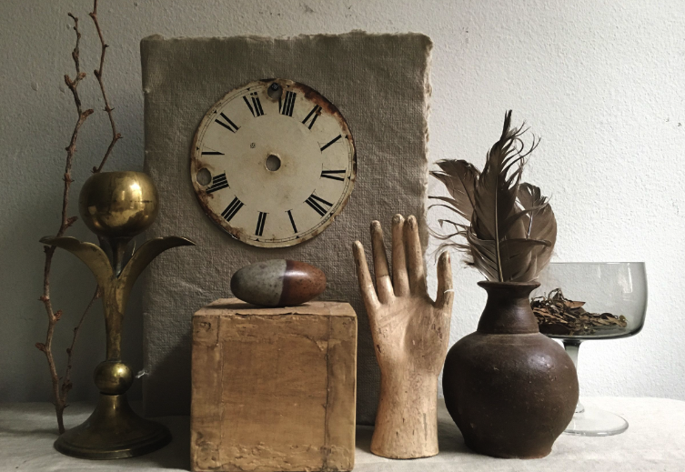 Decorative still life with vintage clock, wooden box, ceramic hand sculpture, vase with feathers, glass bowl, and dried plant on a shelf or table.