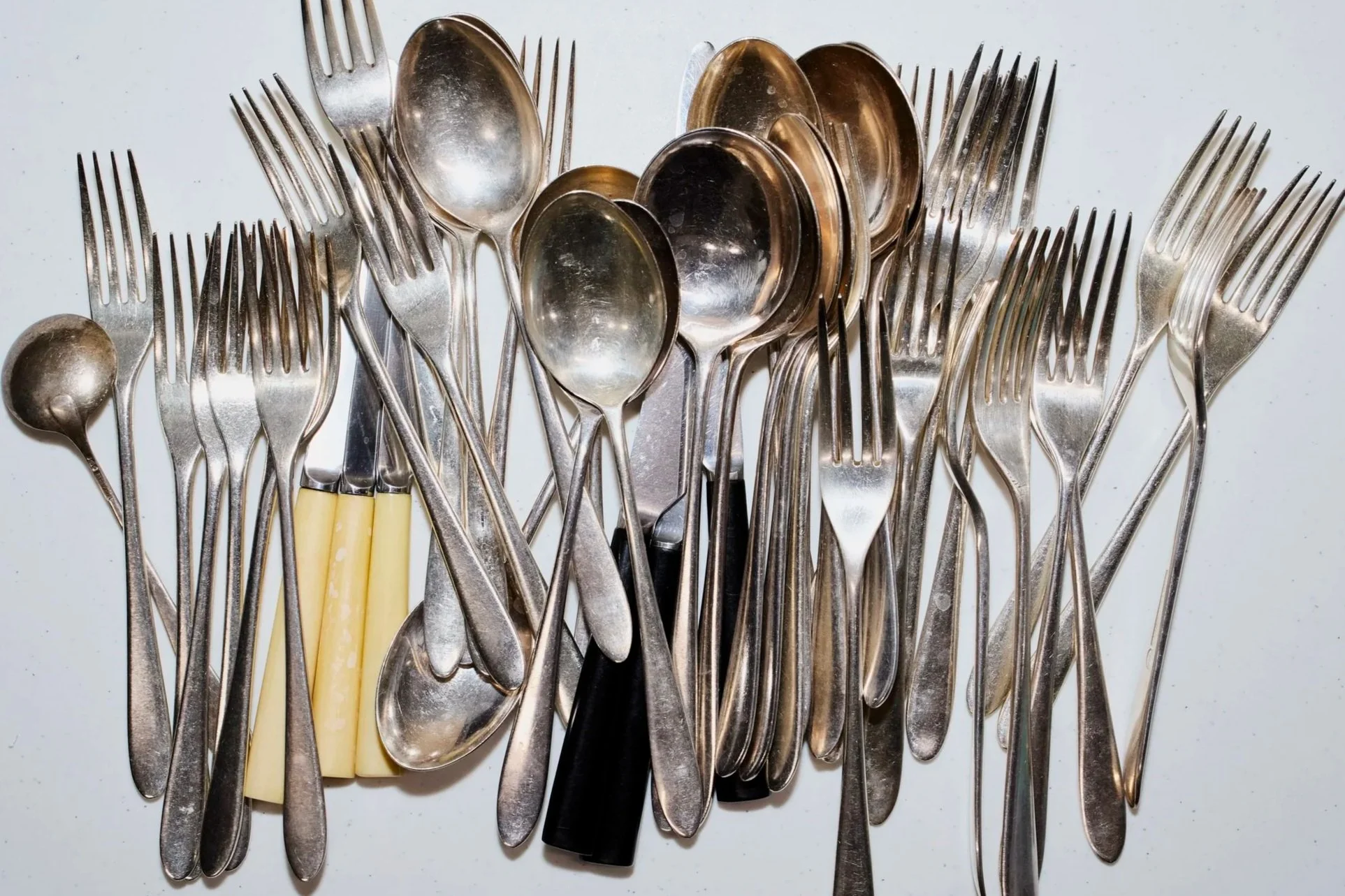 A collection of used silverware, including forks, spoons, and a few knives, arranged on a white surface.