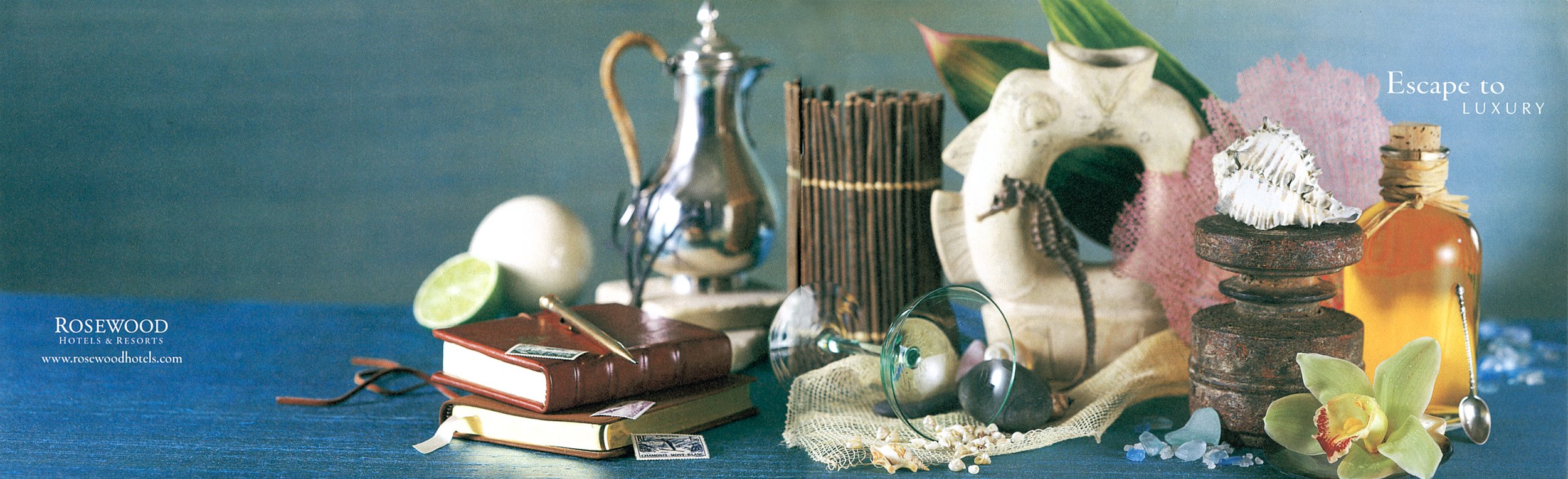 A collection of eclectic decorative items on a blue surface, including a silver teapot, a half lime, a closed red book, a clear glass, seashells, a pink mesh cloth, a large white seashell, a statue of a human skull, a stack of rusted disks, an amber glass bottle, a green orchid flower, and a small spoon, with the words 'Escape to LUXURY' in the top right corner and 'ROSEWOOD HOTELS & RESORTS' with website in the bottom left corner.