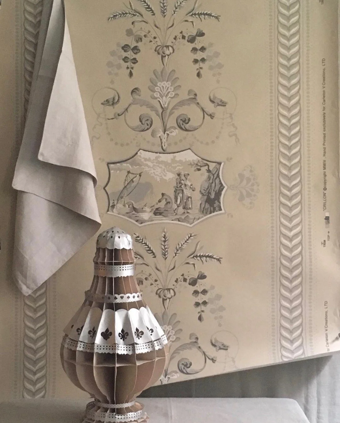A paper table lamp resembling a gourd with intricate cut-out designs and decorative lace ribbons, placed on a white table next to a white cloth napkin, with a vintage wallpaper featuring floral and pastoral motifs in the background.
