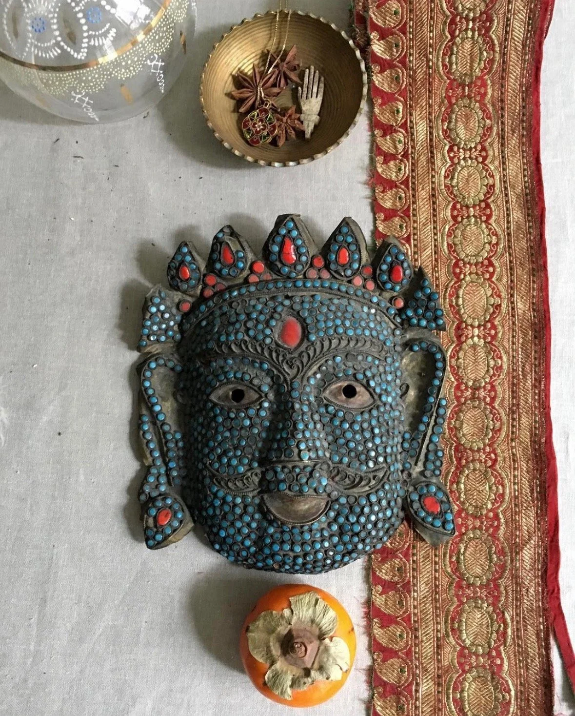 Wall hanging mask of a bearded deity or mythological figure with dark face and blue and red accents, and decorative border textile on the right. Items on the table include a persimmon with a cut stem, a circular dish with star-shaped and wooden objects, and a metallic container with small objects.
