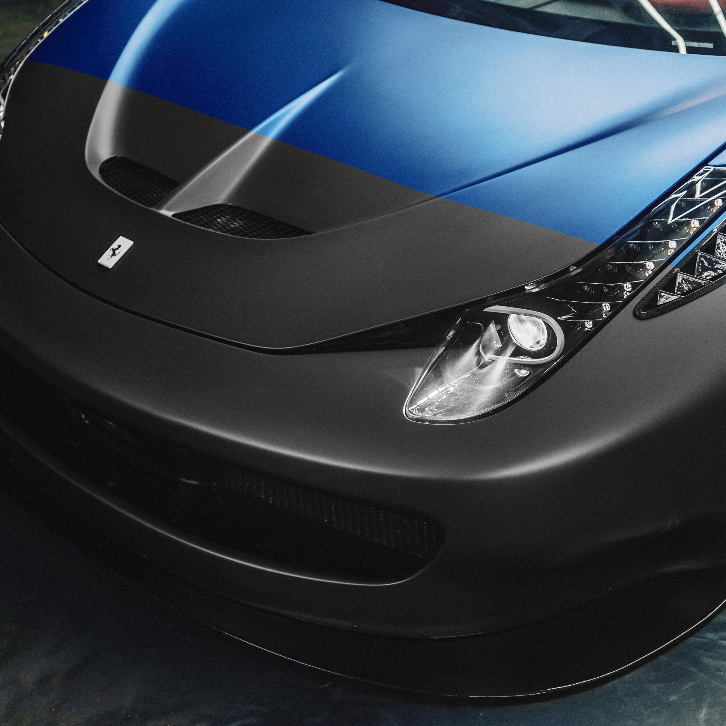 Close-up of a black sports car with a blue racing stripe on the hood, featuring a Ferrari emblem and aerodynamic details.