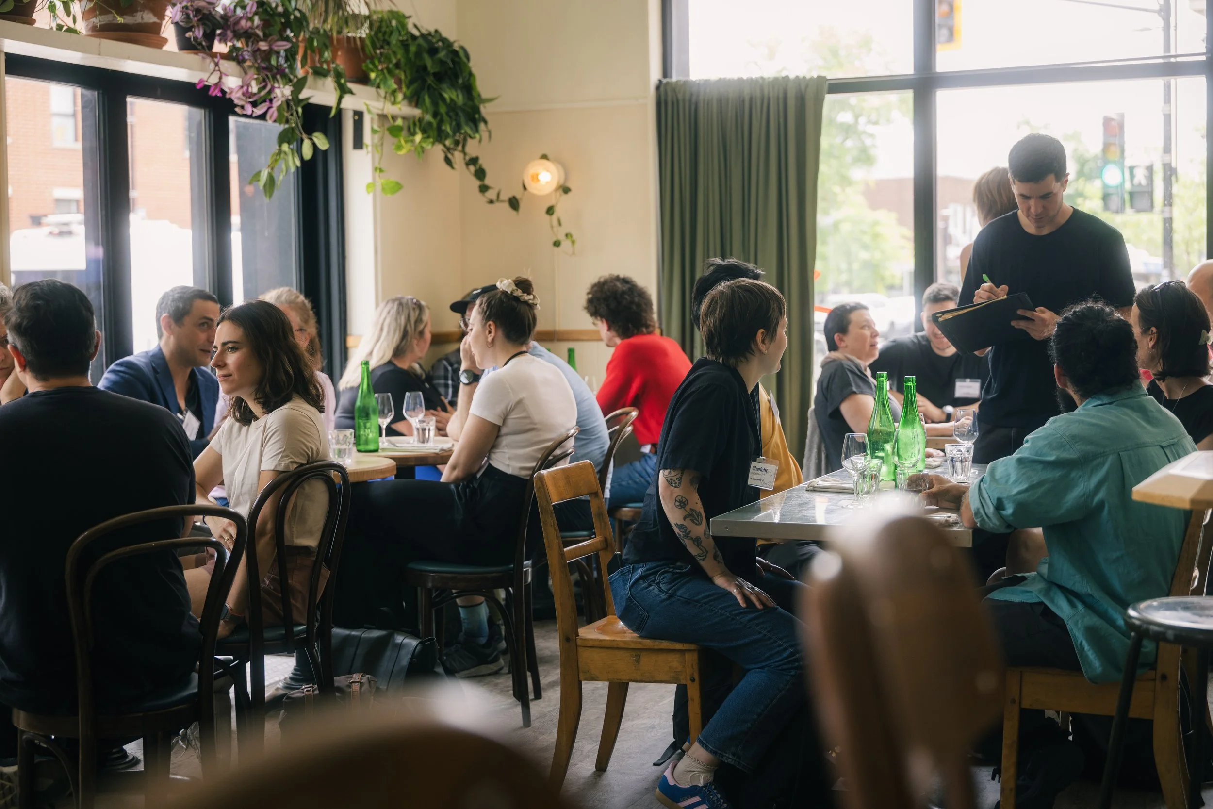 Une salle de restaurant ou de réunion avec plusieurs personnes assises à des tables. Une personne debout semble prendre des notes ou parler, tandis que les autres sont engagés dans des discussions. Beaucoup de bouteilles d'eau vertes sur les tables, décoration végétale près des fenêtres. Ambiance lumineuse avec de grandes fenêtres laissant entrer la lumière naturelle.