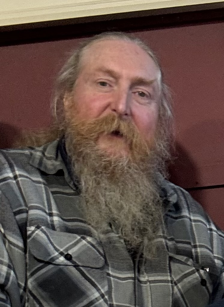 A black and white photo of an older man with a long beard and mustache, wearing a plaid shirt, with a serious expression.
