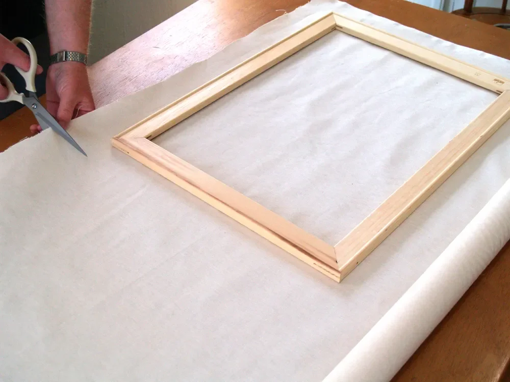 A person cutting a sheet of paper or fabric with scissors near a wooden picture frame on a table.