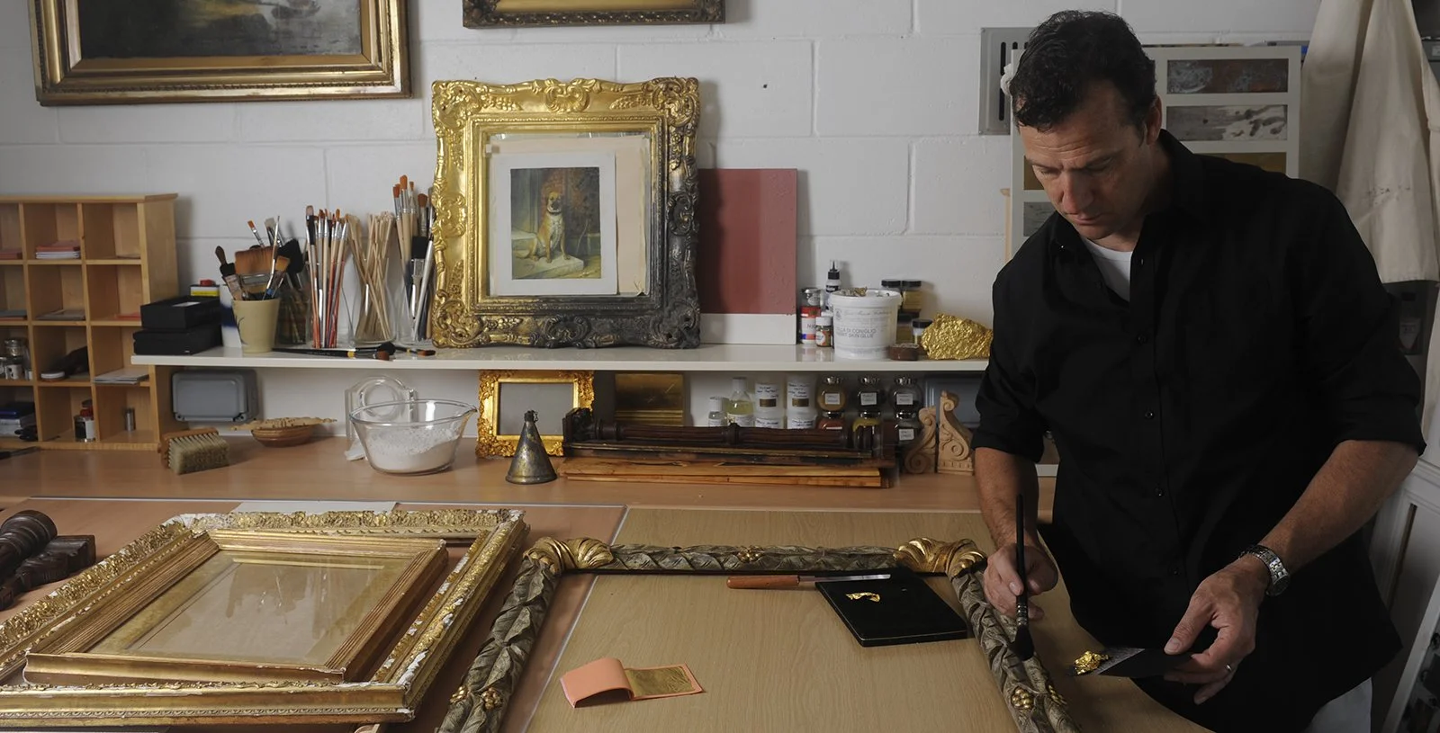 Art conservator working on restoring a gilded picture frame in a studio filled with art supplies, paintings, and decorative objects.