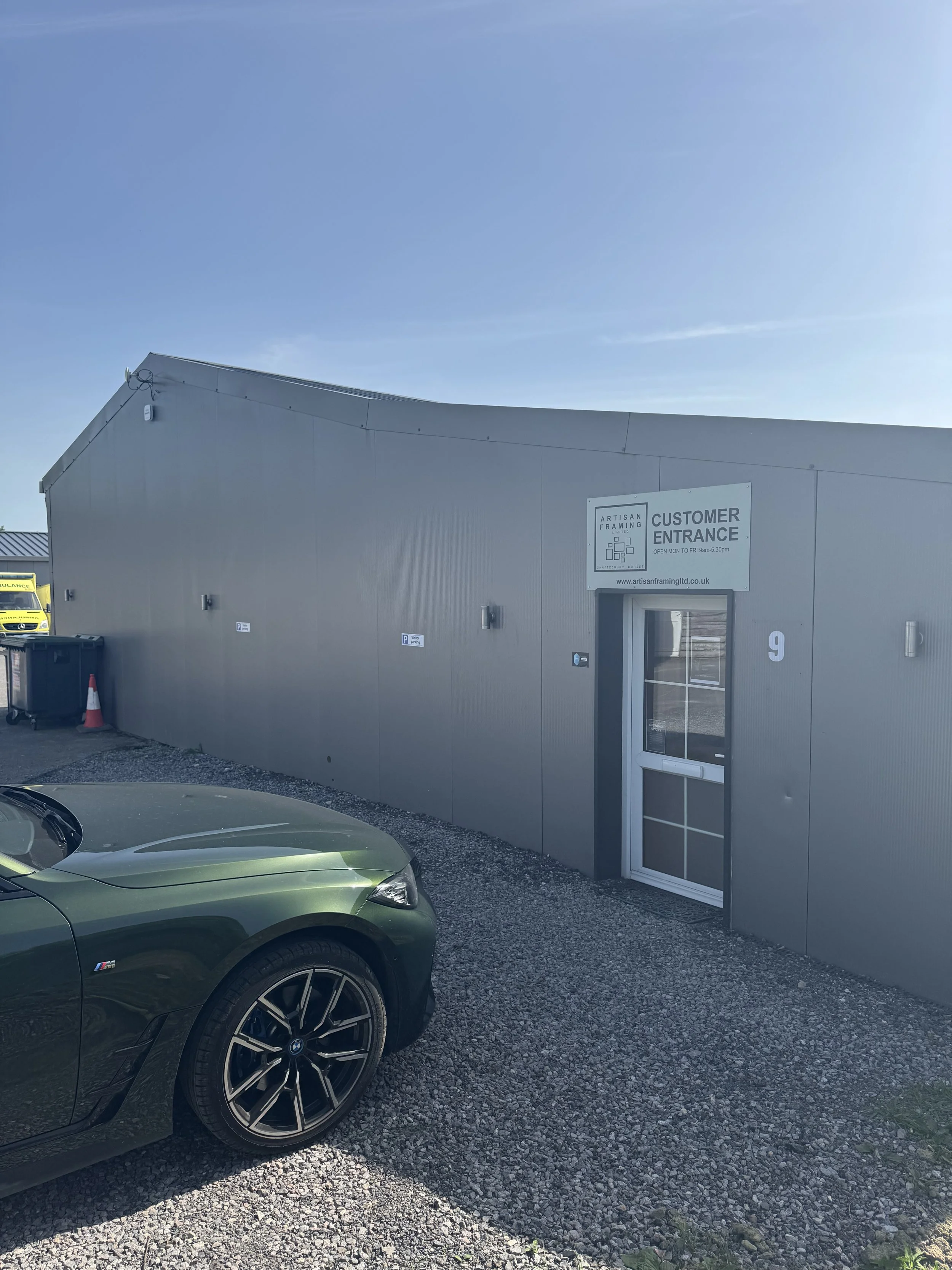 A building with a sign that reads 'Artisan Framing Customer Entrance,' a gray exterior, and a glass door, with a partial view of a green car in the foreground and a yellow ambulance in the background, under a clear blue sky.