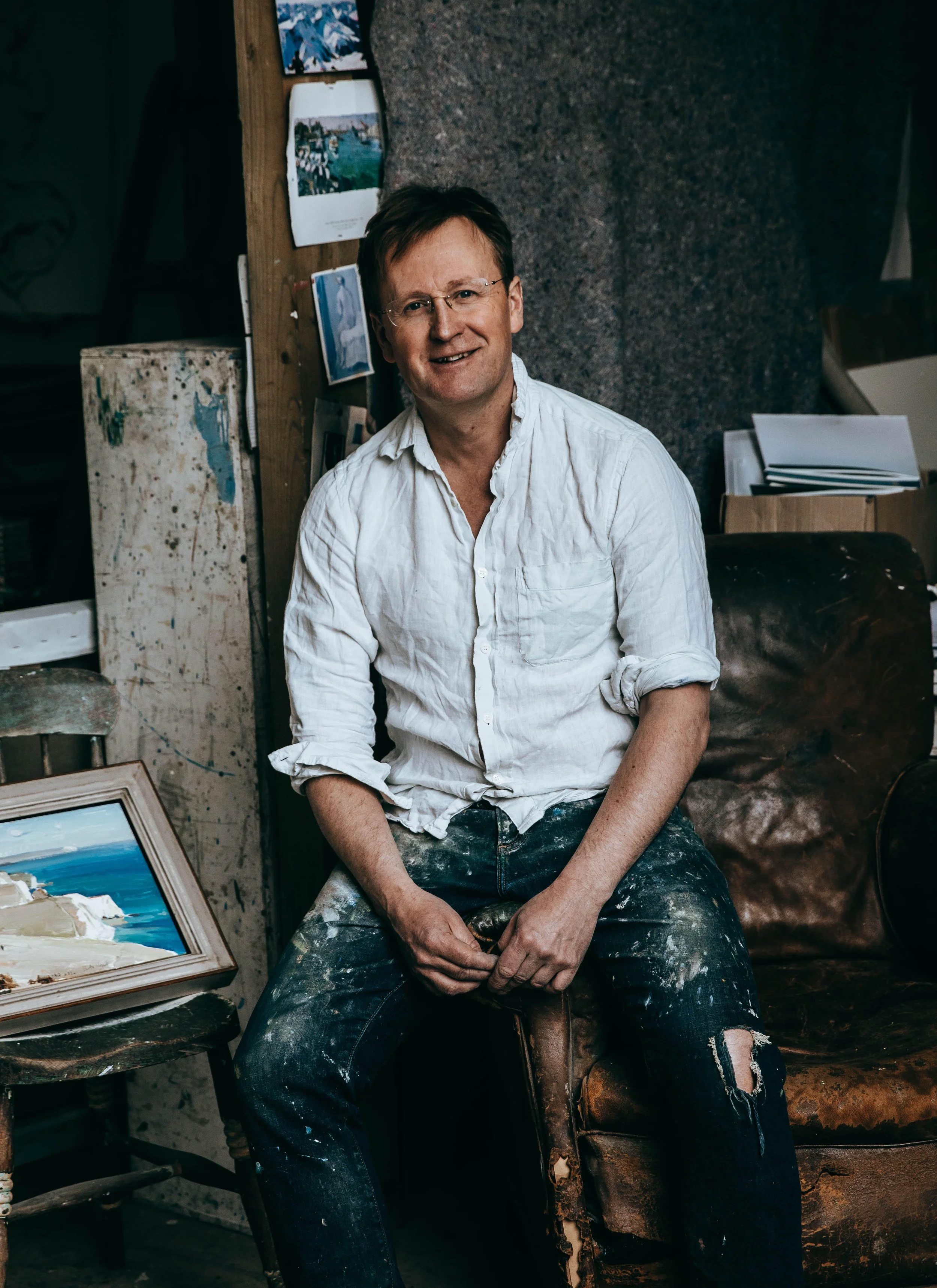 A man with glasses and a white shirt sitting in an art studio, surrounded by art supplies and paintings.