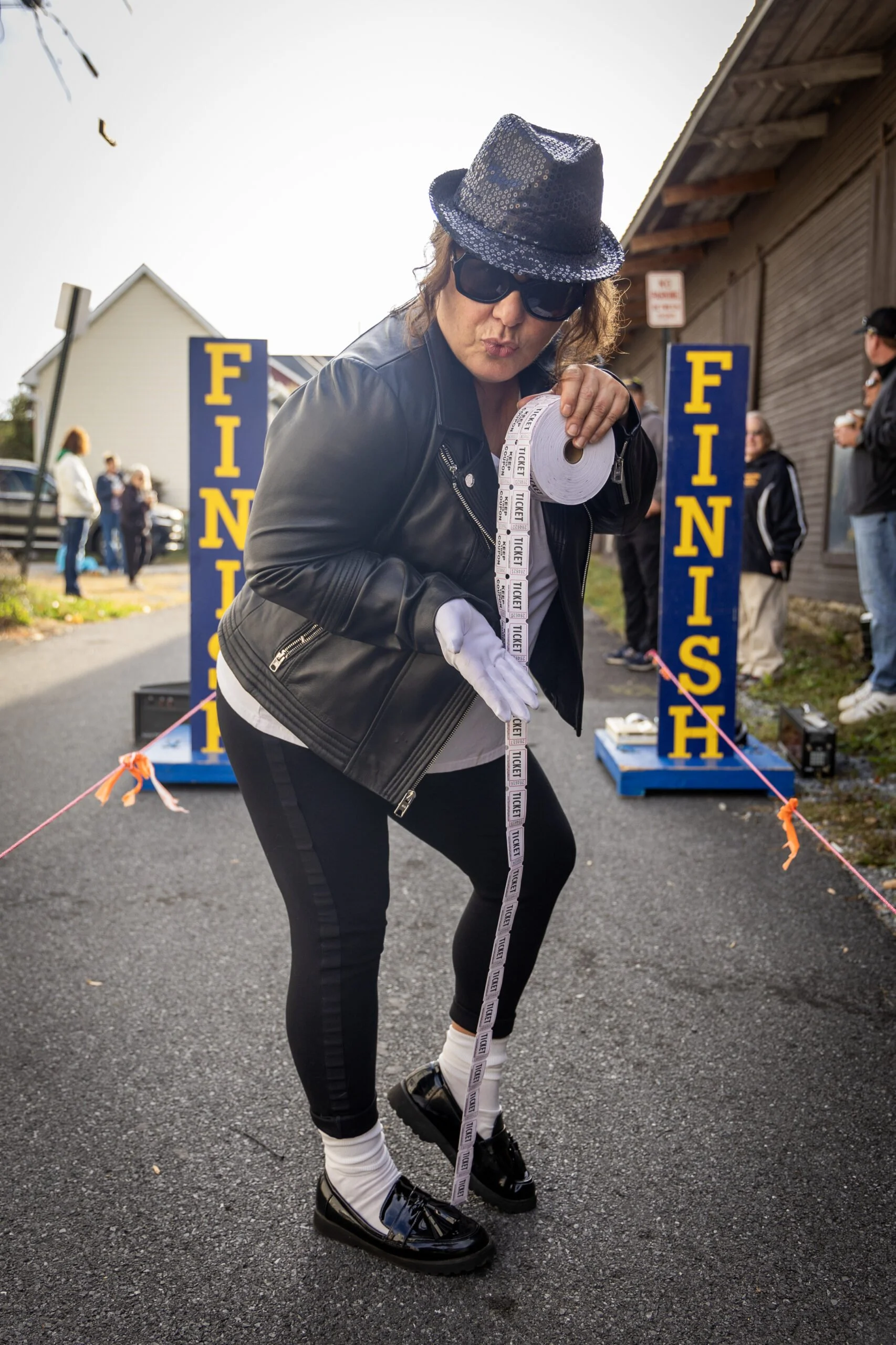 Person dressed in black with a hat and sunglasses at a finish line, holding a roll of tickets and raising a long ticket, with other people and a finish line sign in the background.