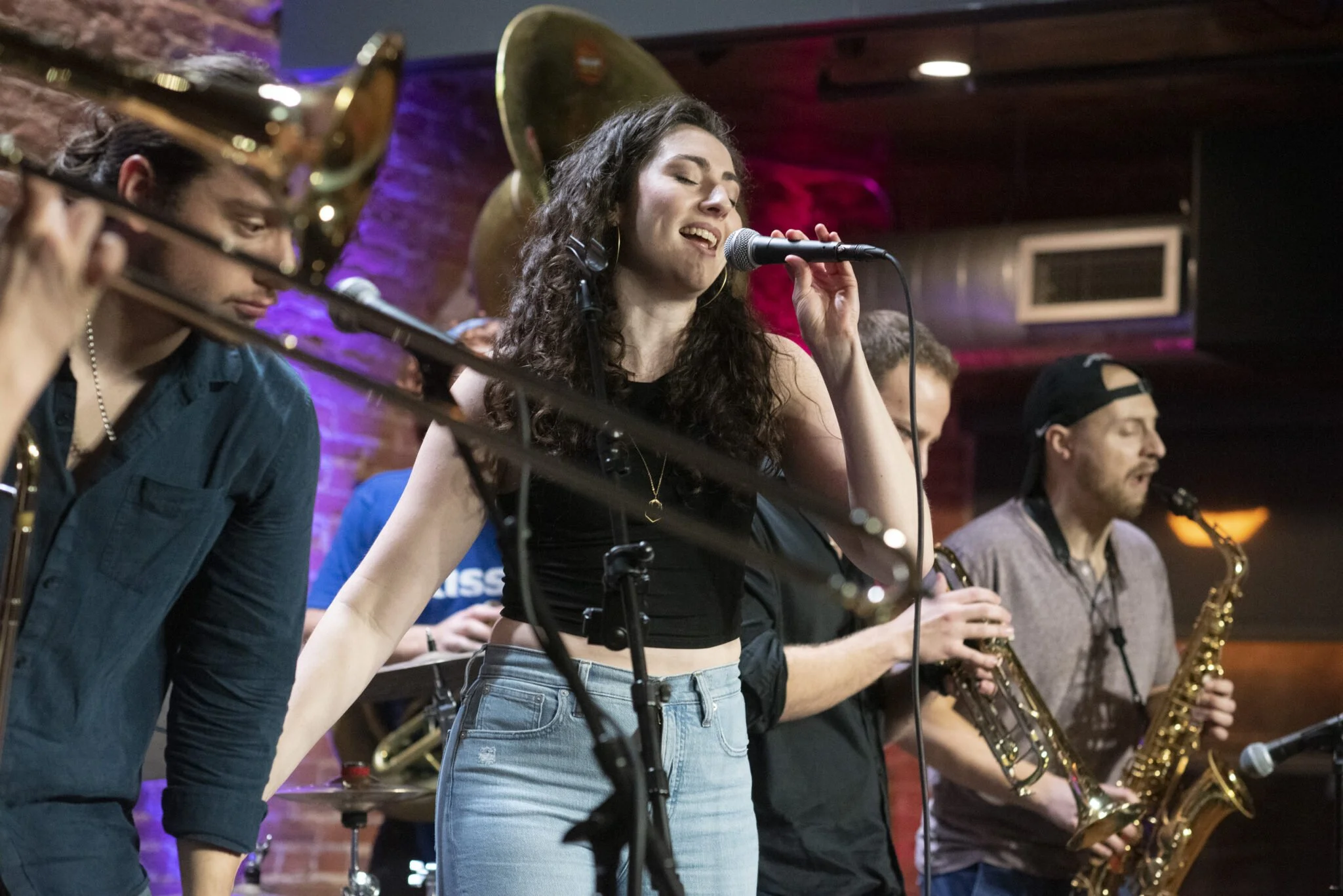 A group of musicians performing live on stage, with a woman singing into a microphone in the center, and two men playing saxophones, in a setting with brick walls and vibrant lighting.
