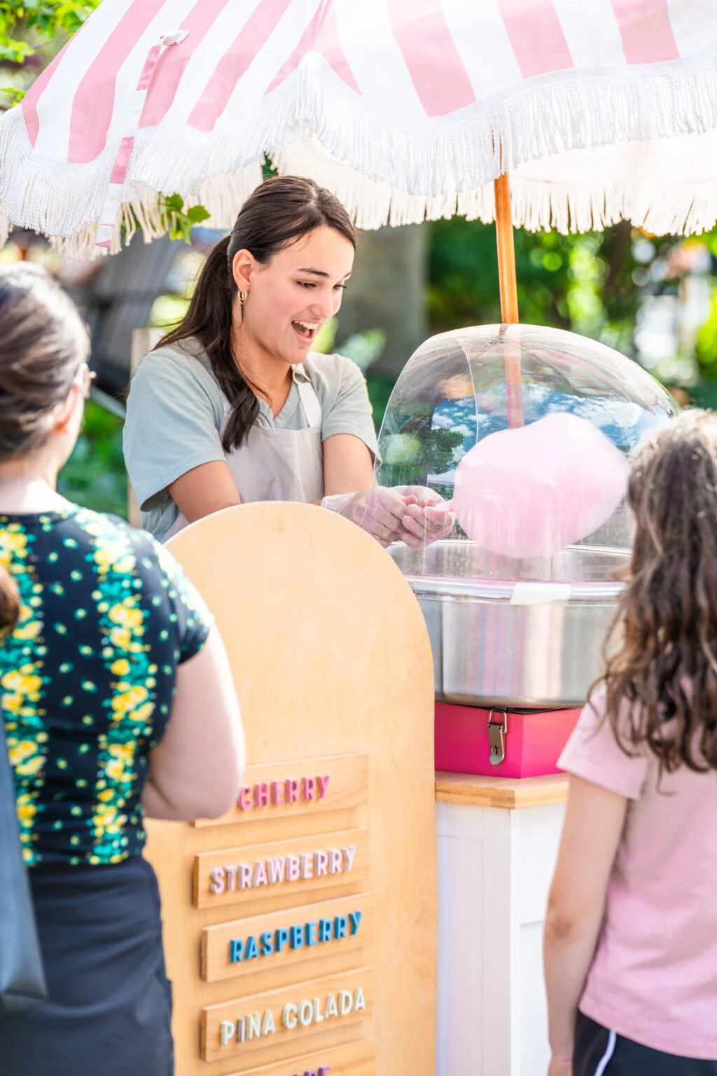 A woman making cotton candy under a pink and white striped umbrella at an outdoor event, with two children watching.