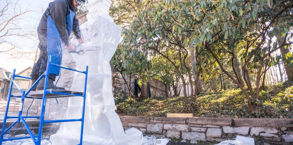 Person using a chainsaw to carve ice sculpture outdoors, standing on a blue scaffold, surrounded by trees and rocks.