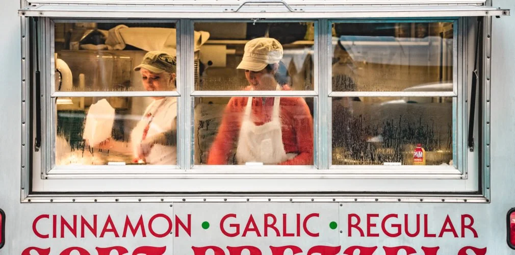 Two women working inside a food truck, seen through a window. One woman wears a beige hat and an apron, the other wears a white cap. The food truck has red and green text advertising cinnamon, garlic, and regular flavors.