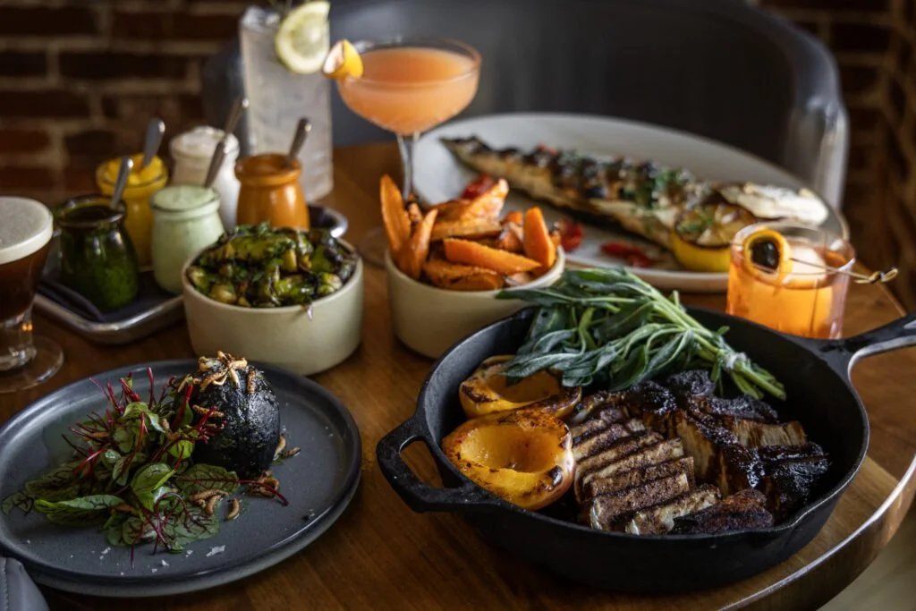A table with a variety of food including grilled meats, roasted vegetables, a whole fish, and sides, along with drinks and condiments.