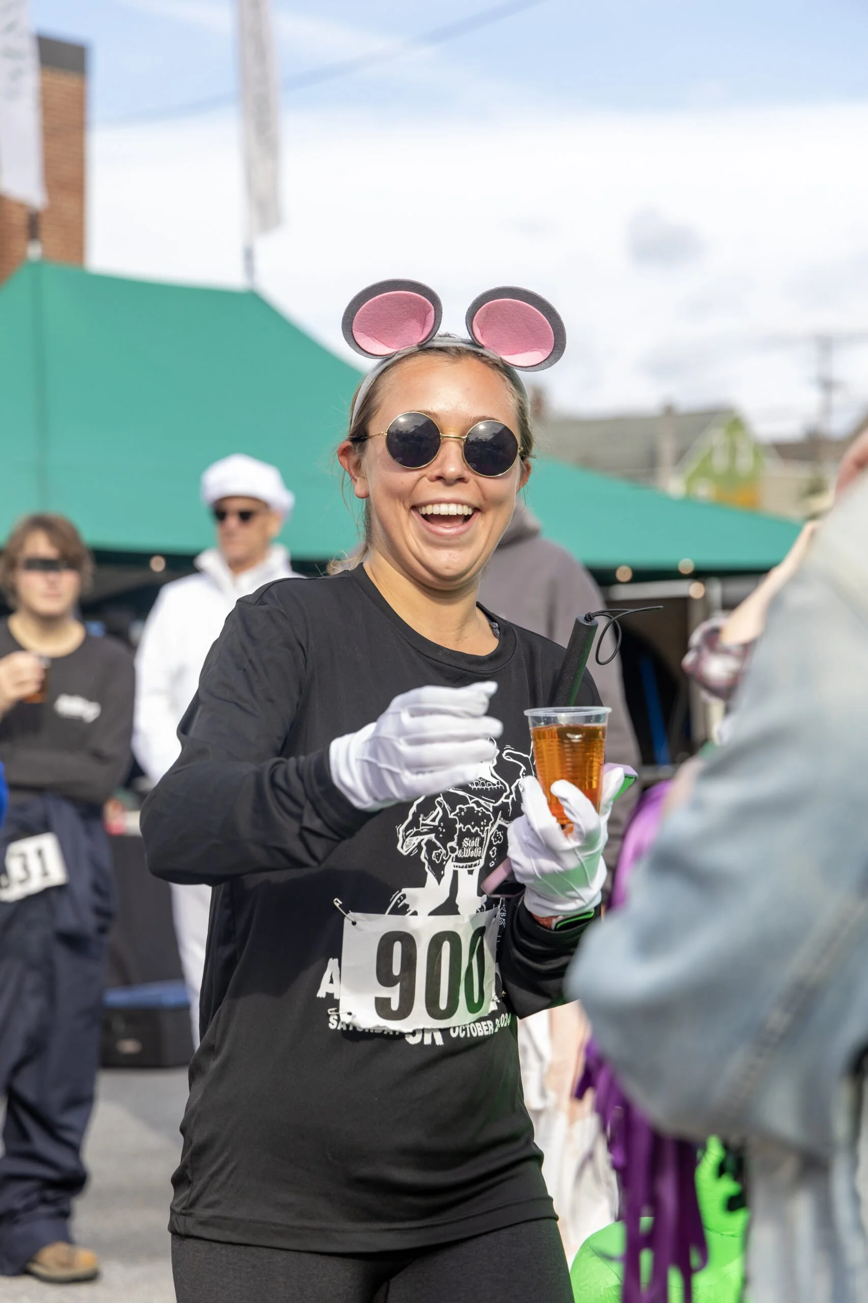 Woman at a race event dressed as a mouse, holding a drink, smiling, wearing sunglasses, mouse ears, black athletic shirt with a race bib number, in a crowded outdoor area with tents and other participants.