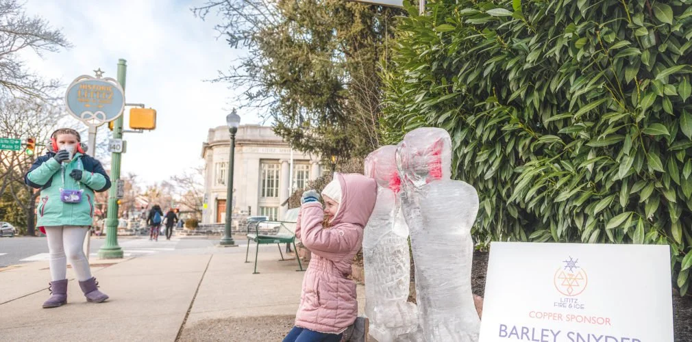 A young girl in a pink coat plays with ice sculptures on a sidewalk in a busy outdoor area, with another girl in a green coat and purple boots nearby.