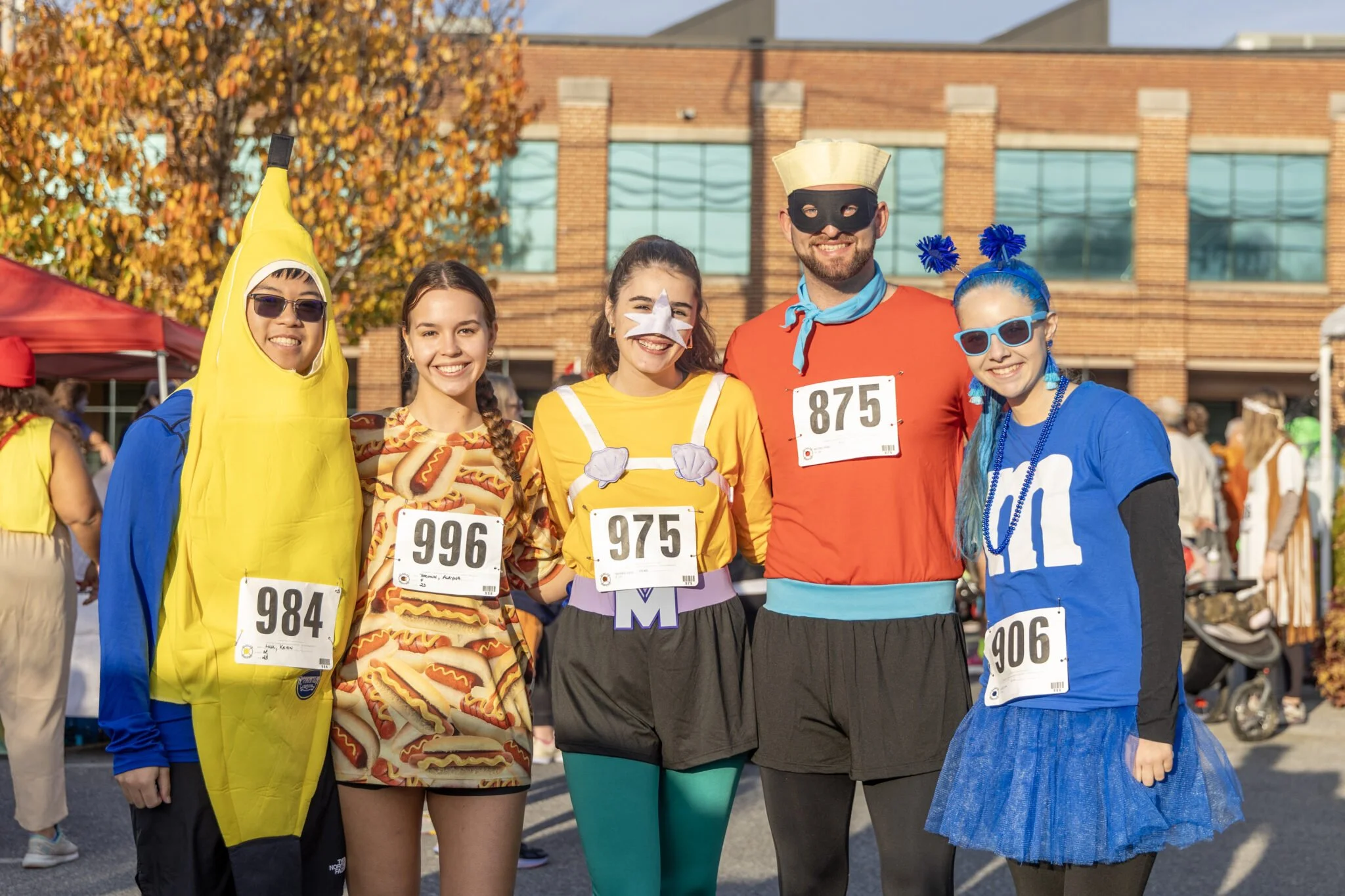 Group of five people dressed in colorful costumes and wearing race bibs, standing outdoors at a fun run event during fall. They are smiling in front of a building with trees displaying autumn leaves.