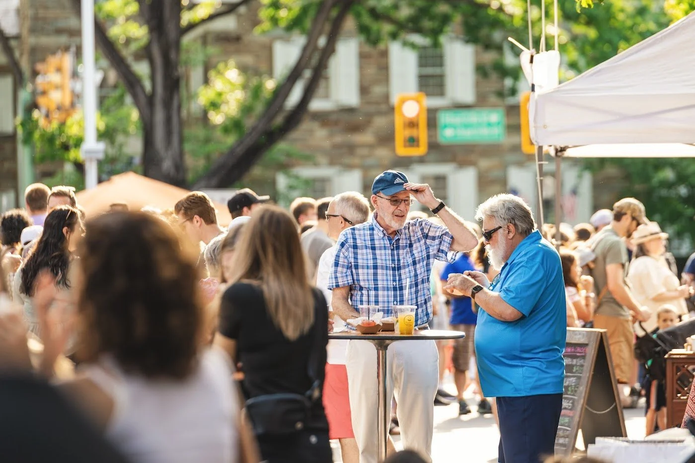 A crowd of people at an outdoor event on a sunny day, with two elderly men in the foreground talking, one in a blue plaid shirt and the other in a blue polo, surrounded by food and drinks.