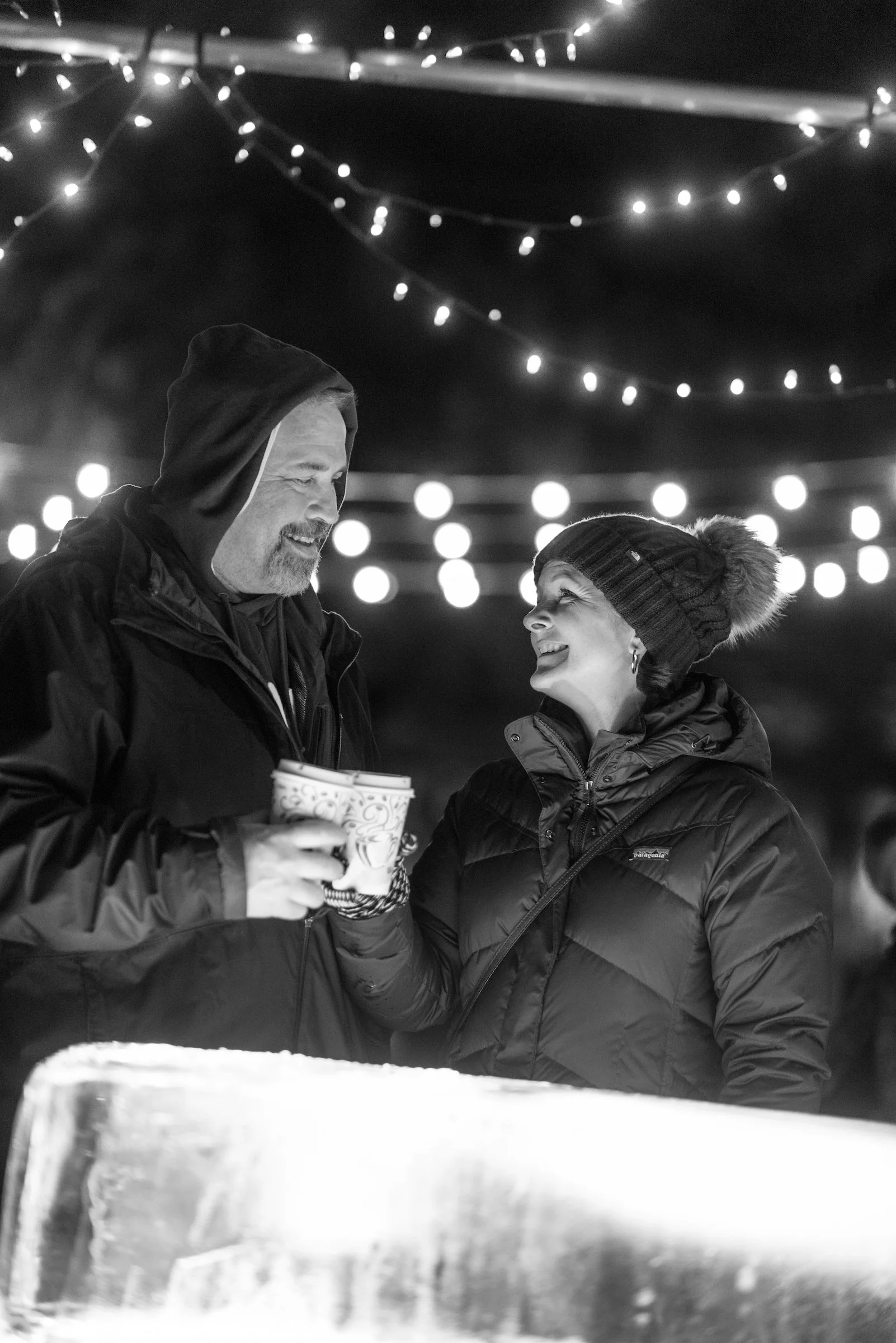 A man and woman in winter clothing smiling at each other while holding cups at an outdoor winter event decorated with string lights.